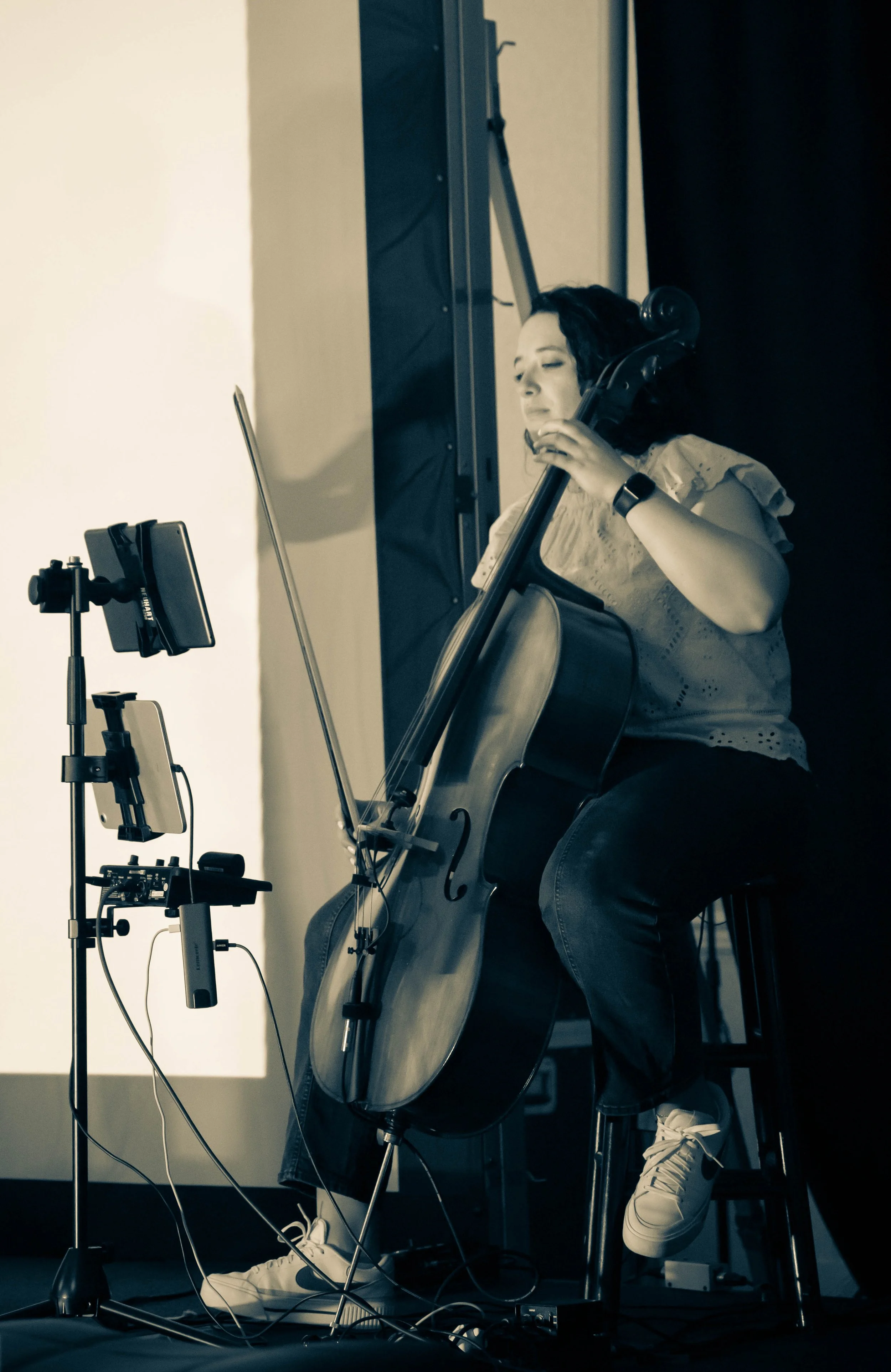 Woman playing a cello with a phone on a stand recording her, wearing a watch and sneakers.