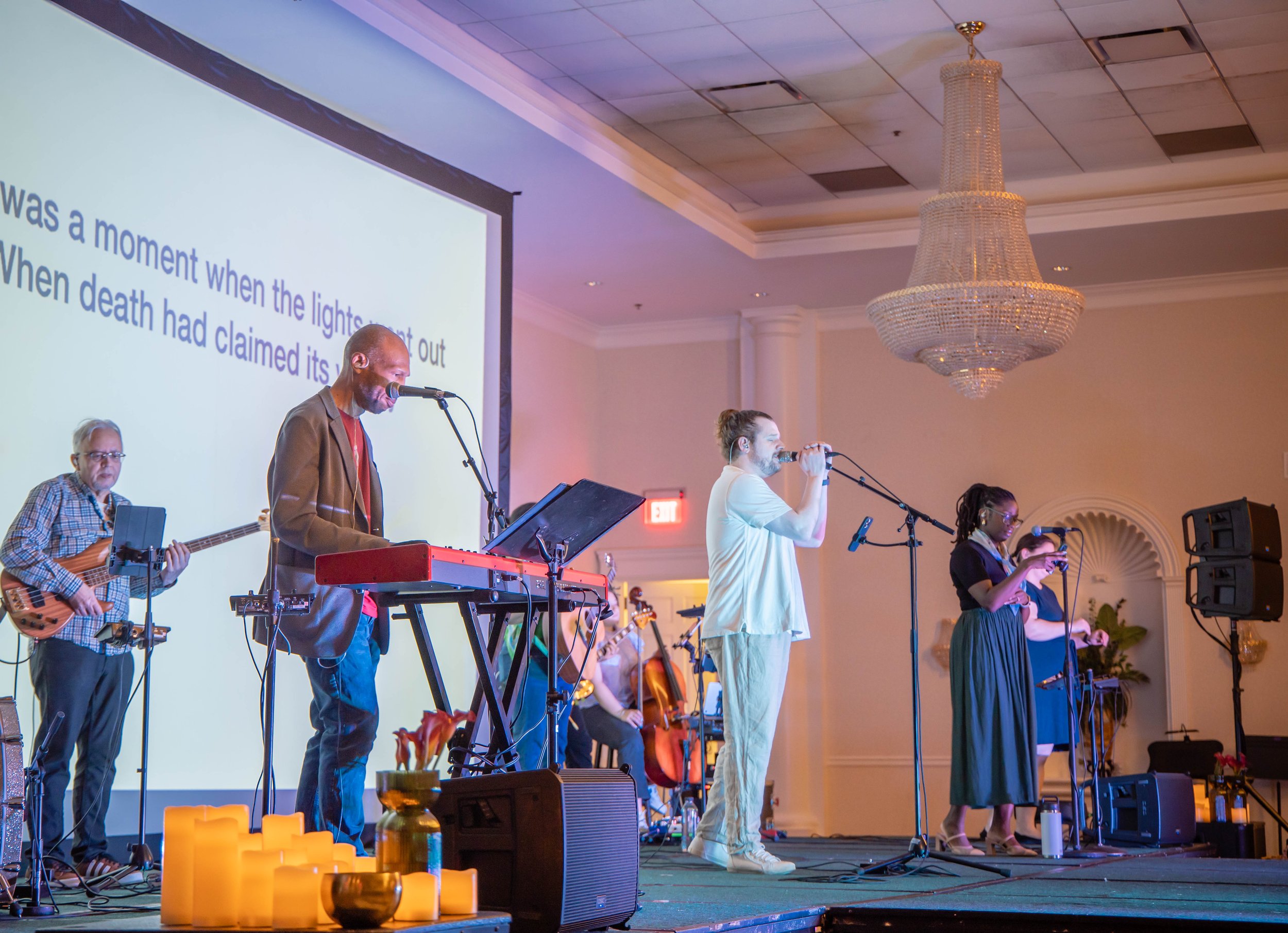 A musical band performing on stage in a banquet hall, with four band members playing instruments and singing into microphones, a large chandelier hanging from the ceiling, and a projection screen in the background displaying song lyrics.