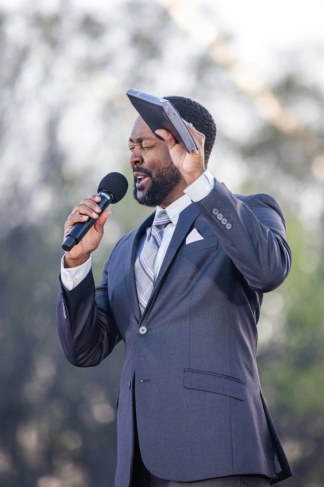 A man in a suit passionately singing into a microphone with a tablet in his hand, outdoors, with a blurred natural background.