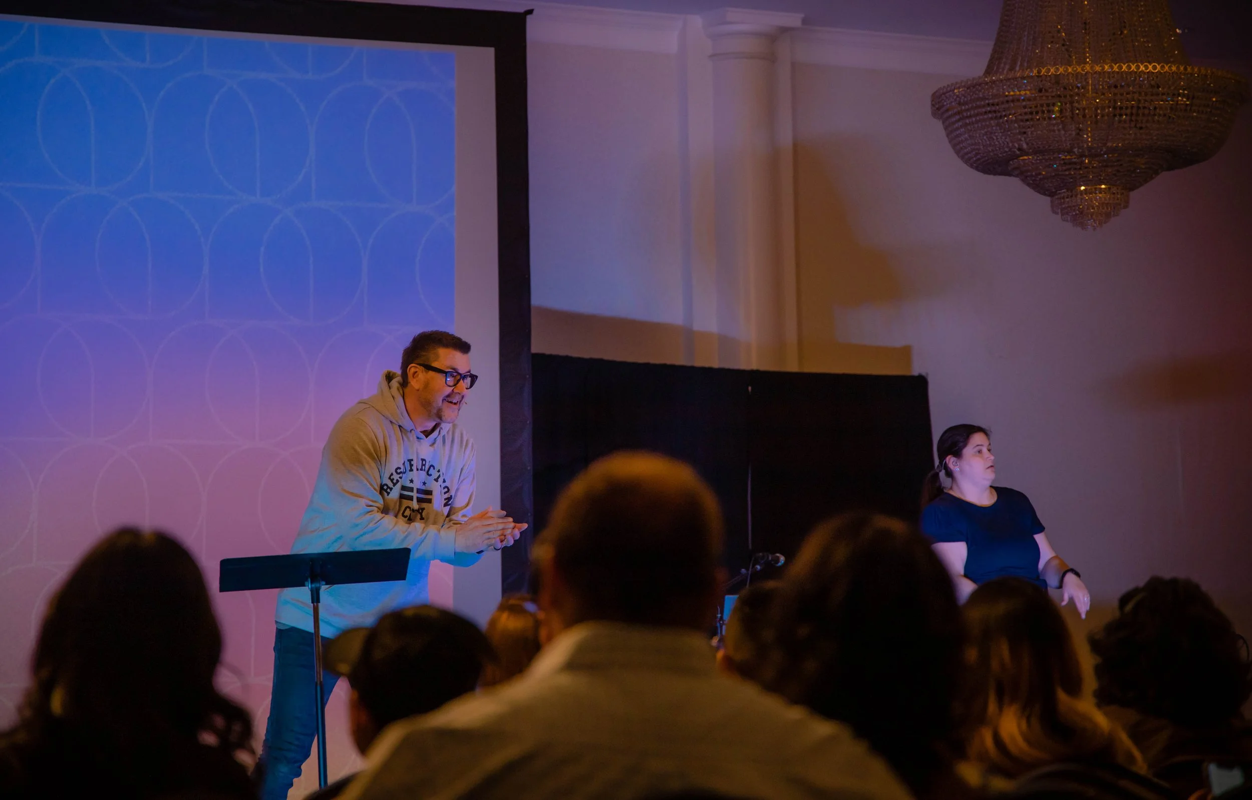 A man and a woman standing on a stage in front of an audience, with a large projection screen behind them. The man is wearing glasses and a hoodie, and appears to be speaking or presenting, while the woman is standing with her arms crossed, listening