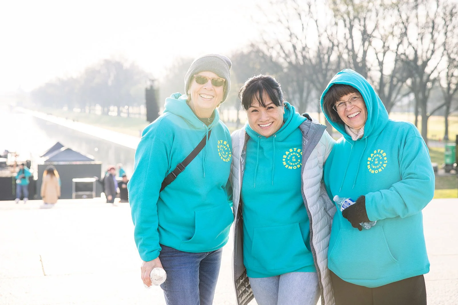 Three women wearing matching light blue hoodies with yellow text, standing outdoors on a bright day, smiling for the camera.