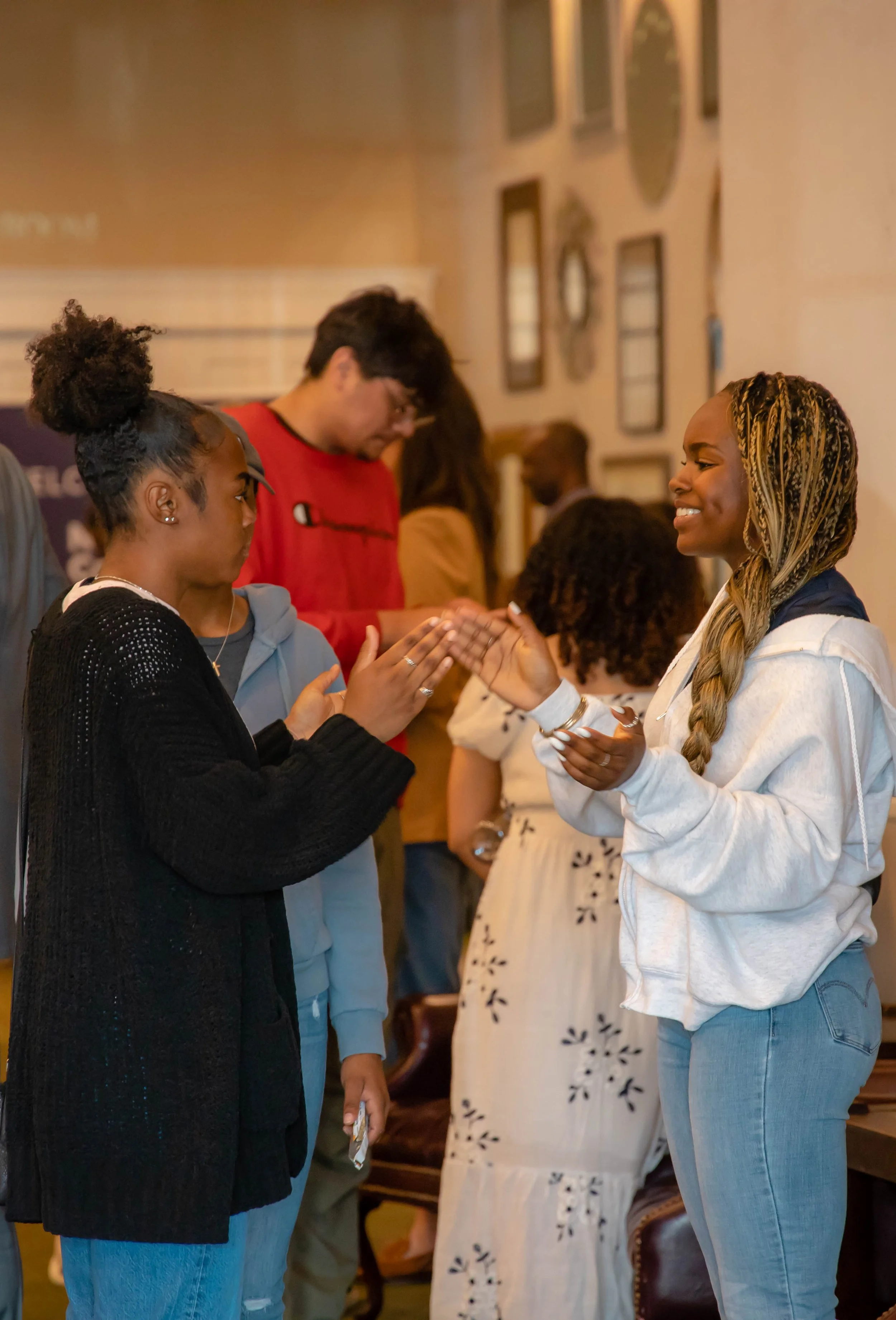 Two women are exchanging a handshake while talking. Several people are standing in line behind them in an indoor setting decorated with wall art.