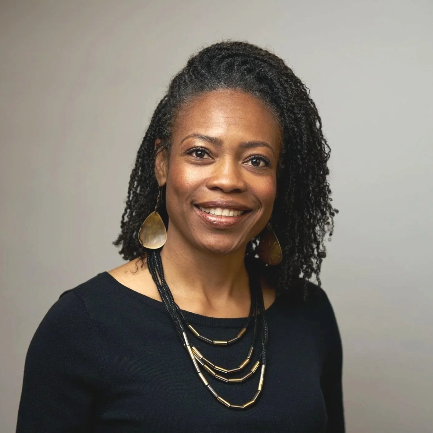 A smiling woman with black hair in twists with black and gold necklace and gold earrings.
