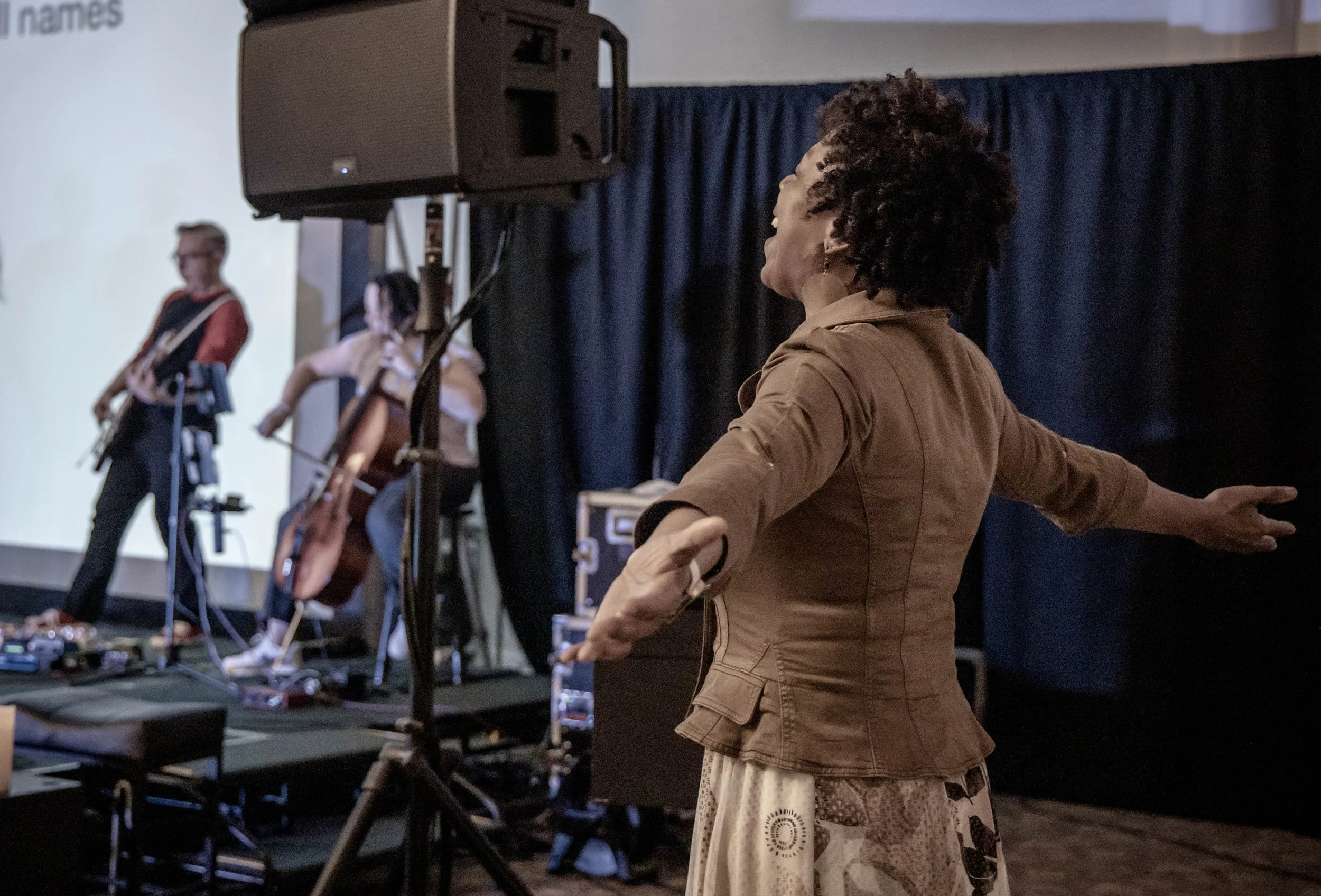 A woman with curly hair dancing or singing with her arms outstretched, performing on stage with a band in the background.