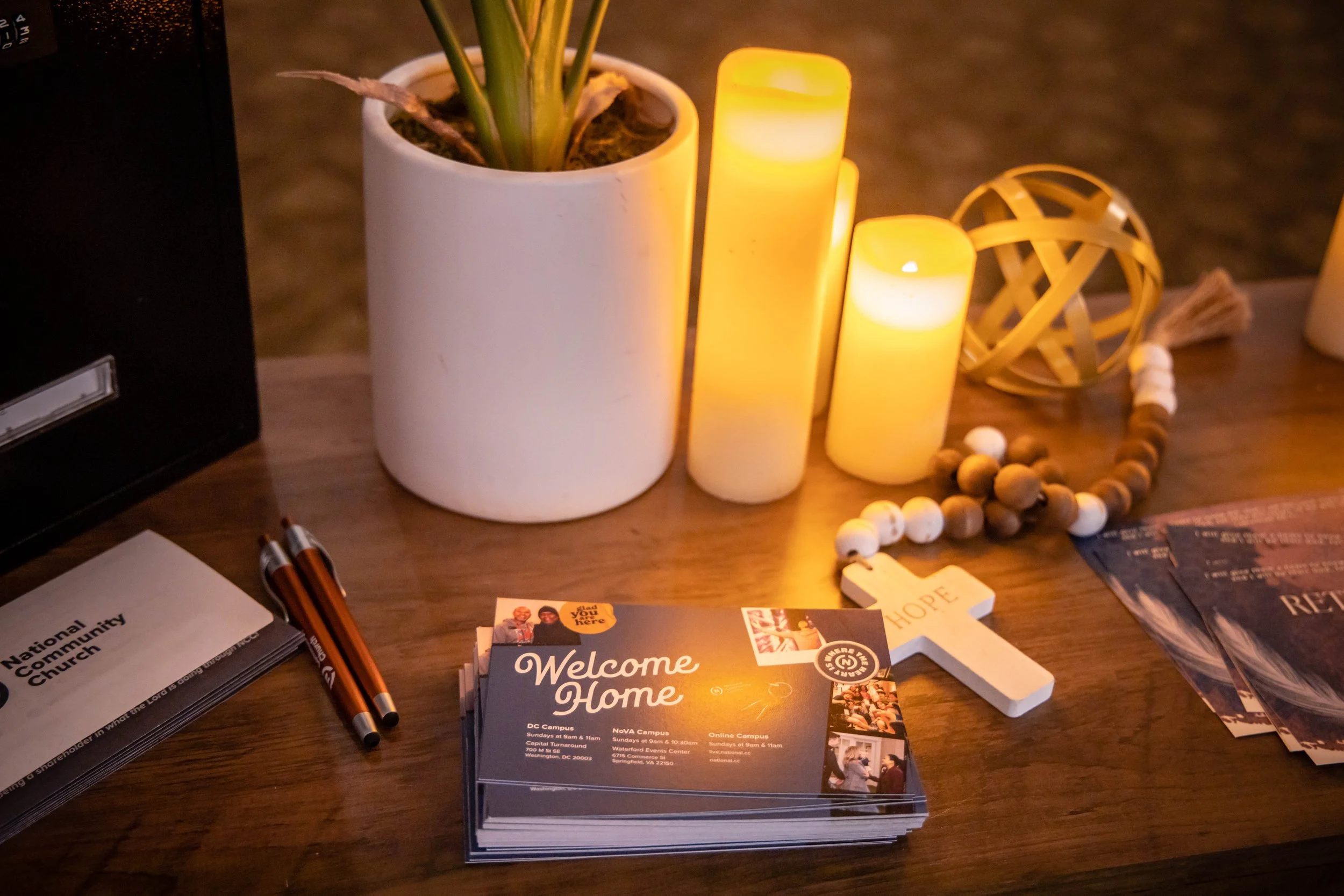 A wooden table with a white potted plant, three yellow candles, prayer beads, a white cross, brochures, and pens.