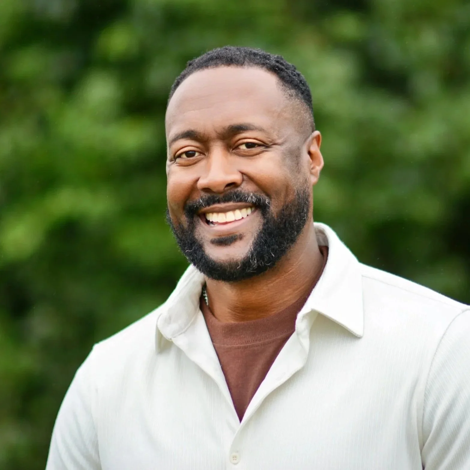 A smiling man in a white collared shirt, in front of greenery outdoors