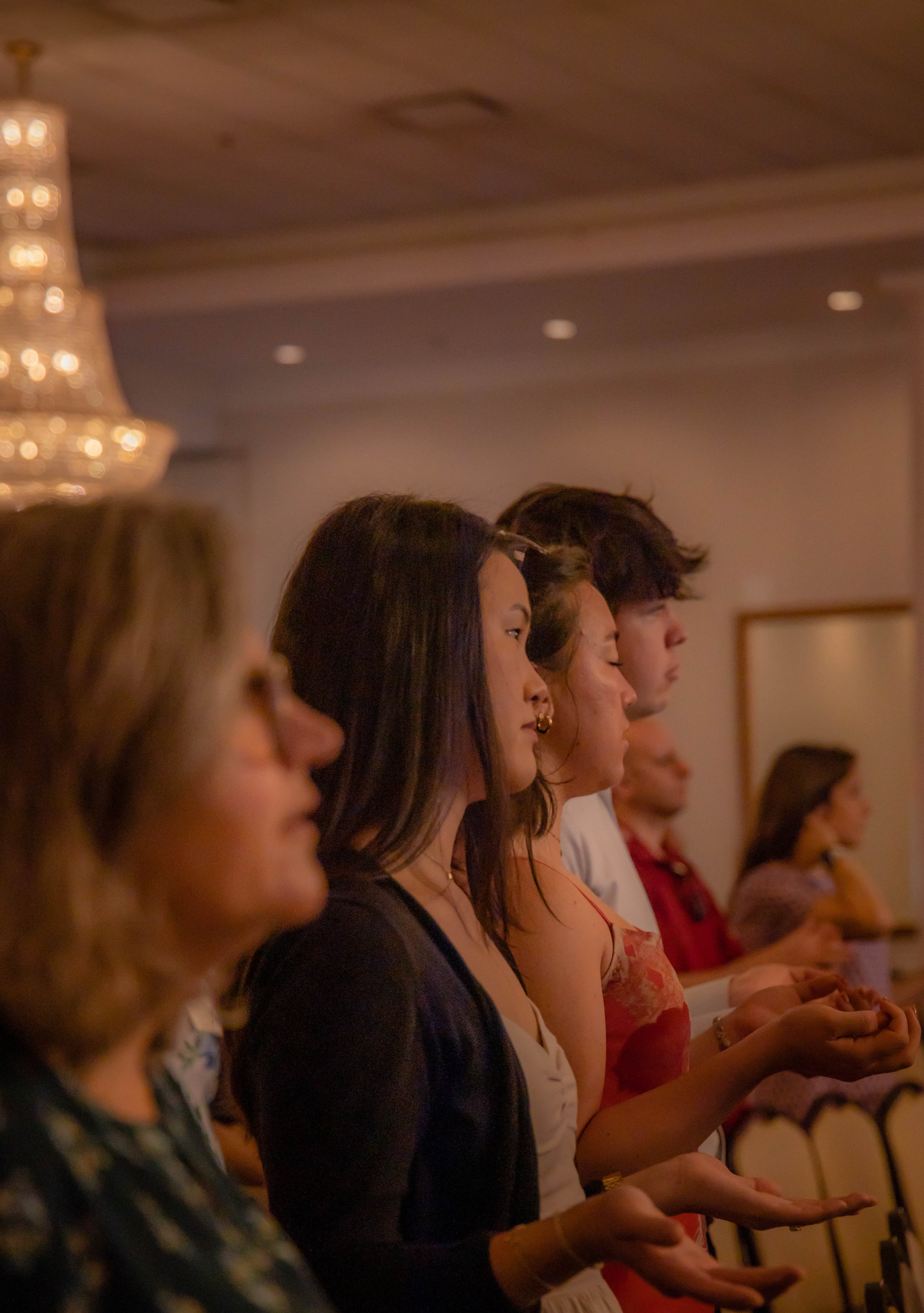 A line of people standing with their eyes closed, meditating or praying in a dimly lit indoor setting, with a chandelier visible in the background.
