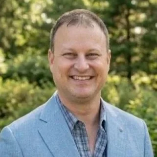 Smiling man in a plaid button up shirt with blazer with greenery in background.