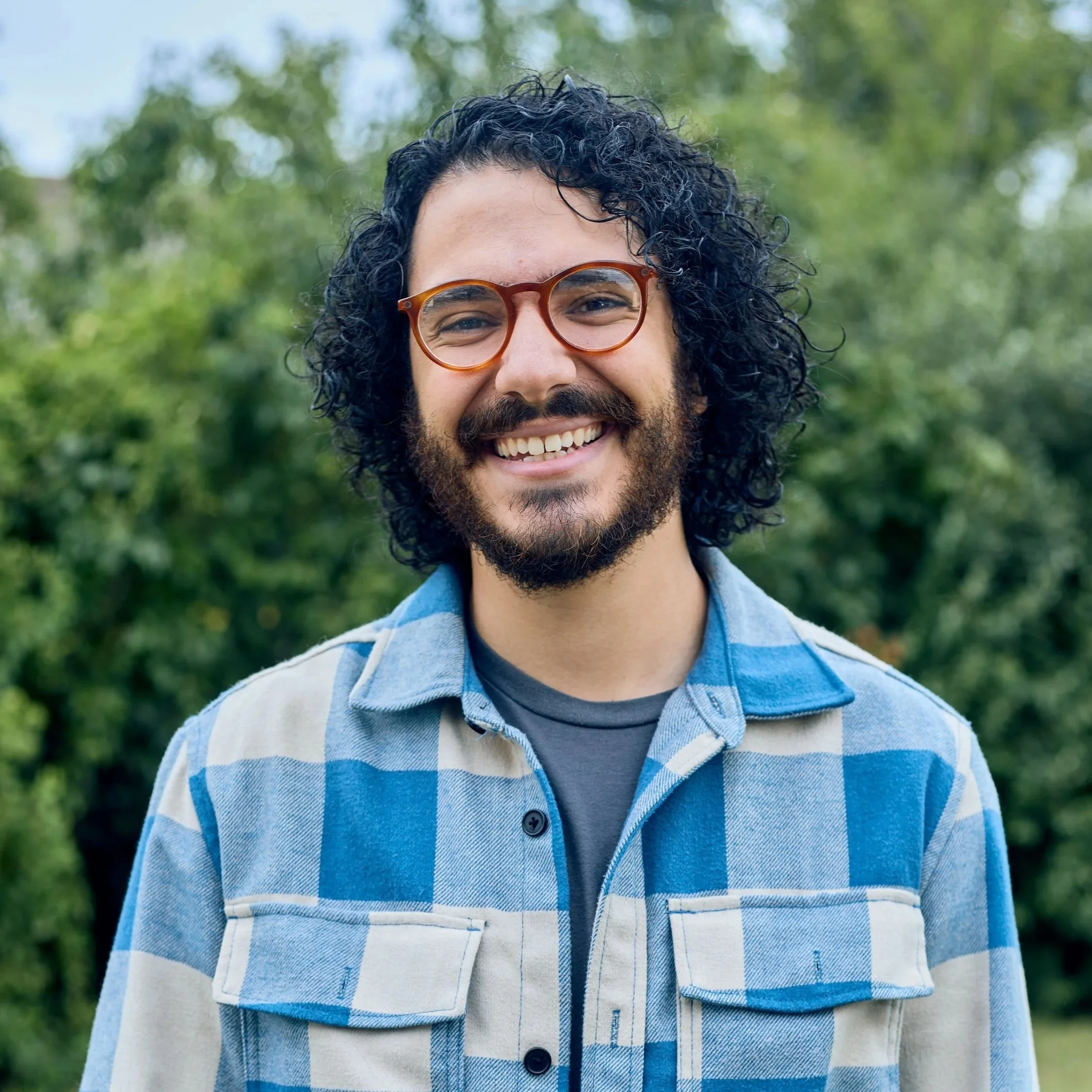A smiling man with curly dark hair and a beard, wearing glasses with orange frames, standing outdoors with blurry green and brown background.