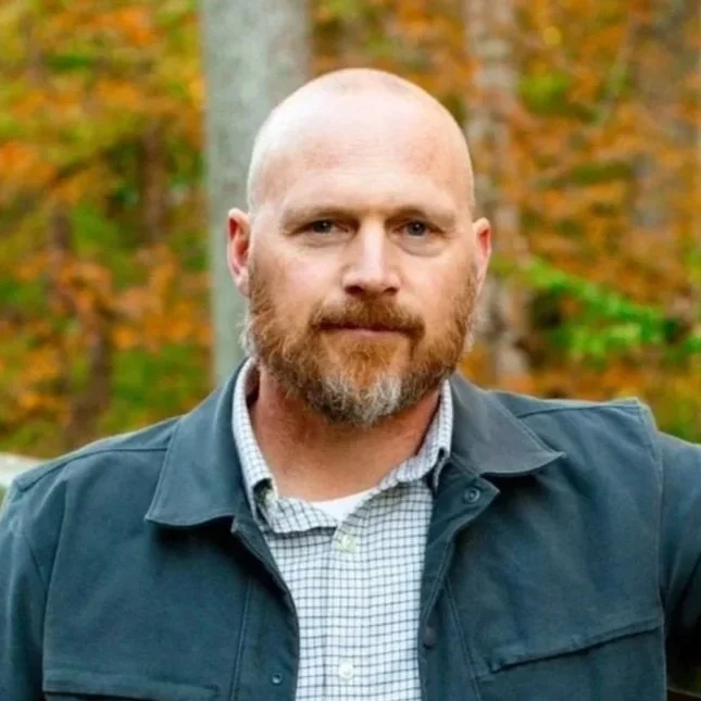 A man with a beard outside on a bridge with Autumn trees behind him.