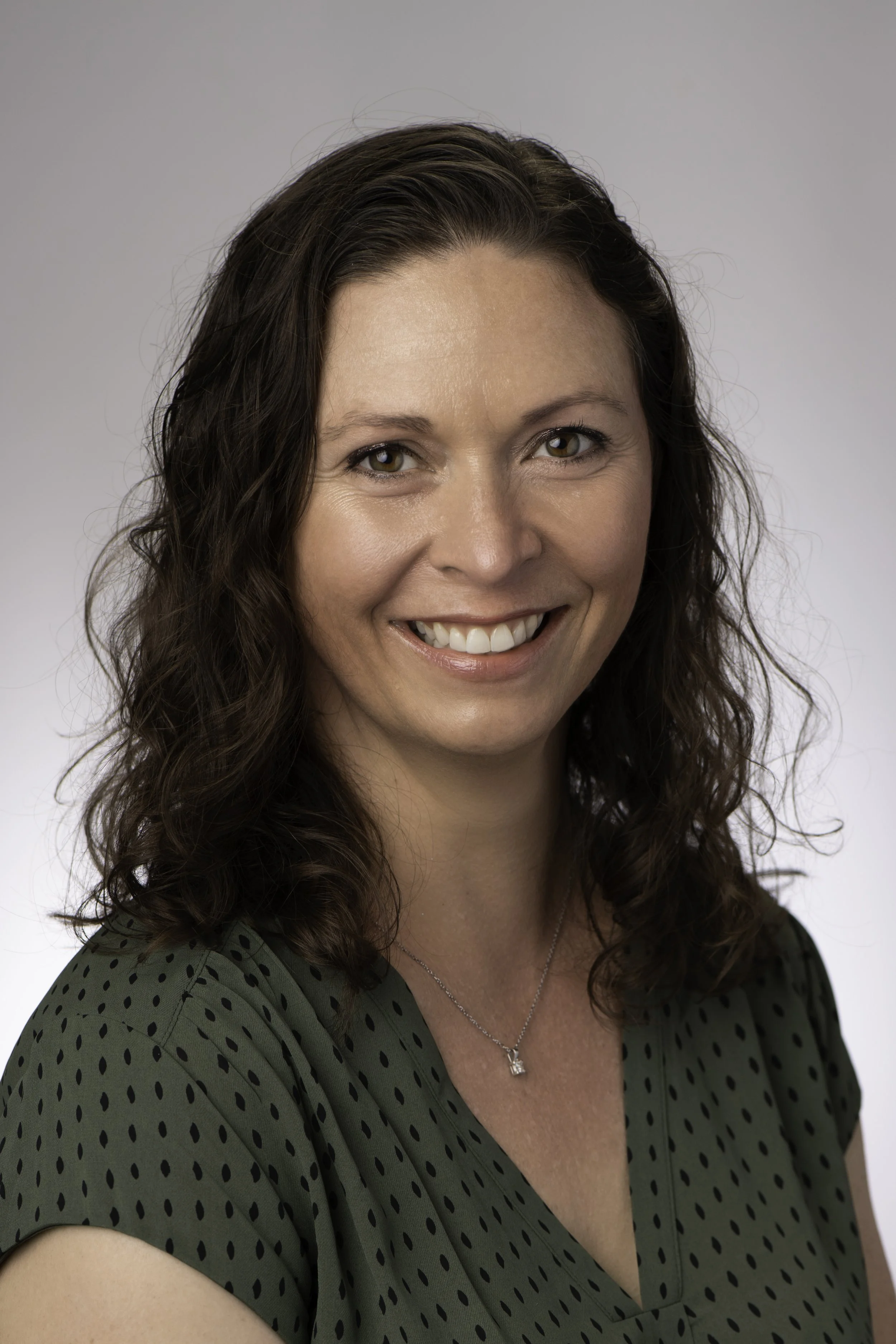 A woman with shoulder-length curly brown hair, smiling, wearing a green blouse with black polka dots and a silver necklace, against a plain gray background.