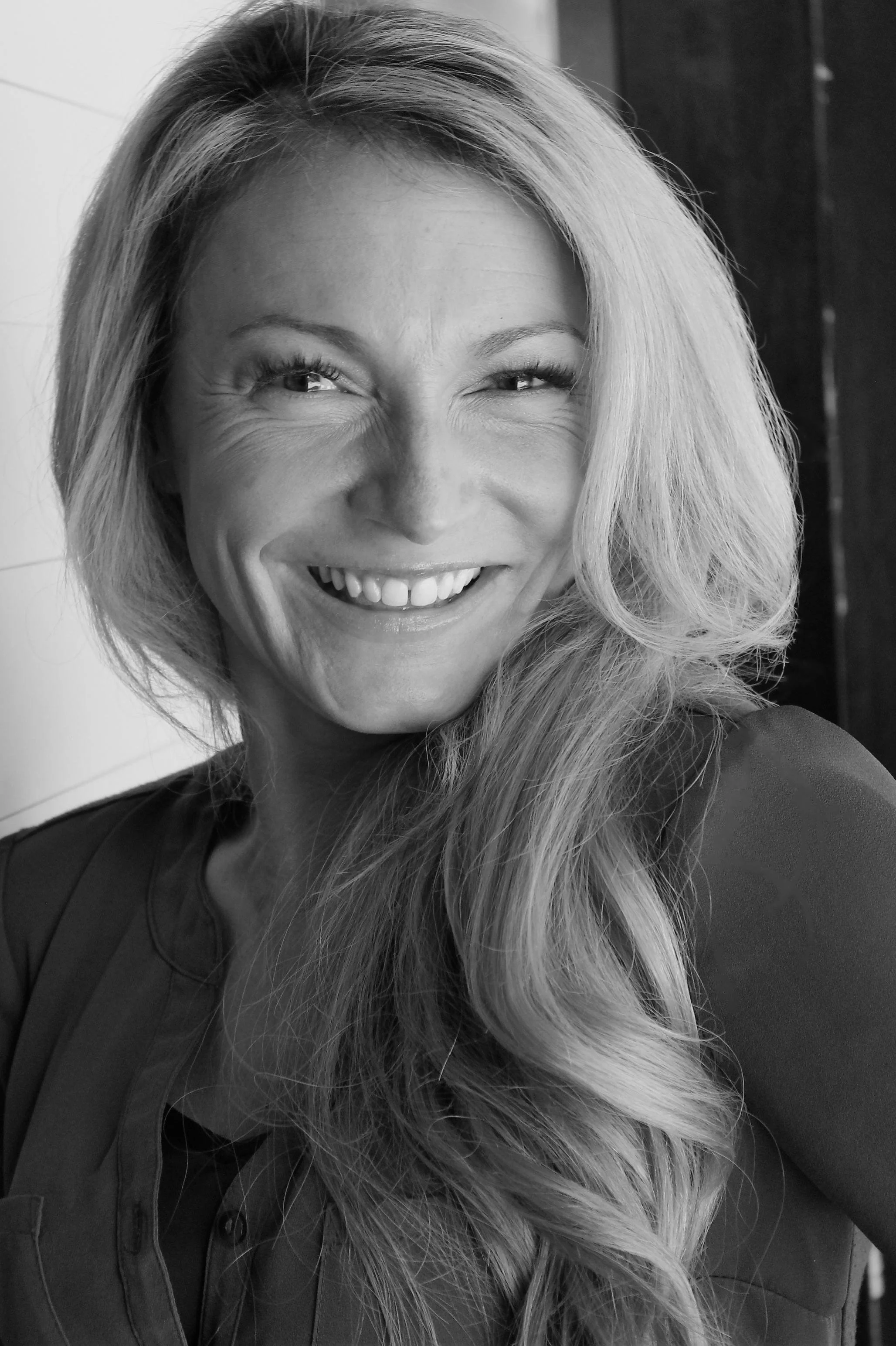 Black and white photo of a woman with long, wavy hair smiling and squinting with one eye, showing her teeth.