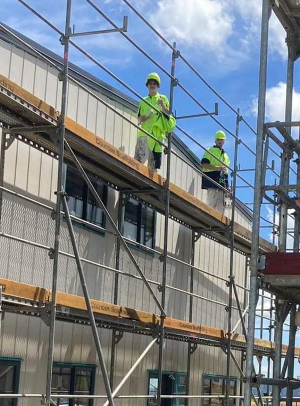 Two construction workers in neon safety vests and helmets standing on scaffolding outside a building under a blue sky with white clouds.