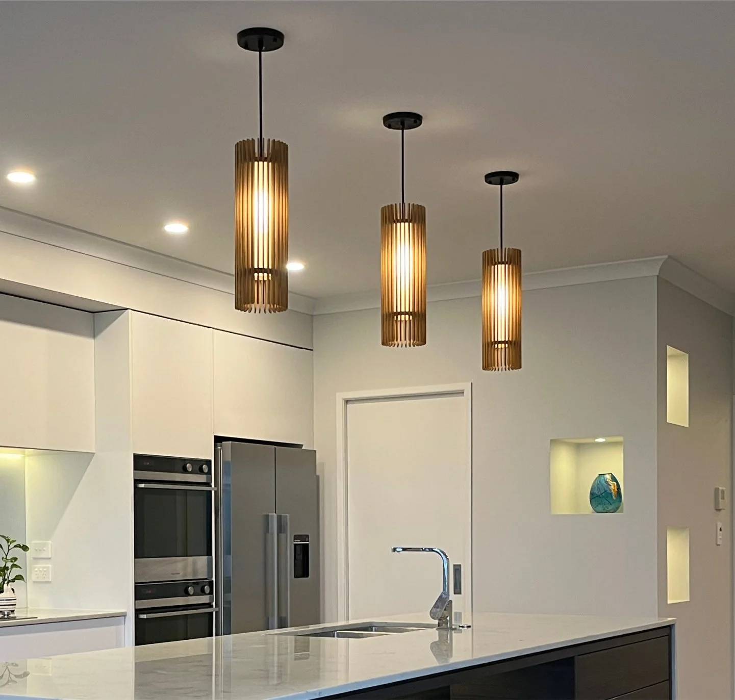 Modern kitchen with three hanging wooden pendant lights, stainless steel refrigerator, built-in oven, white cabinetry, and an island with a sink.