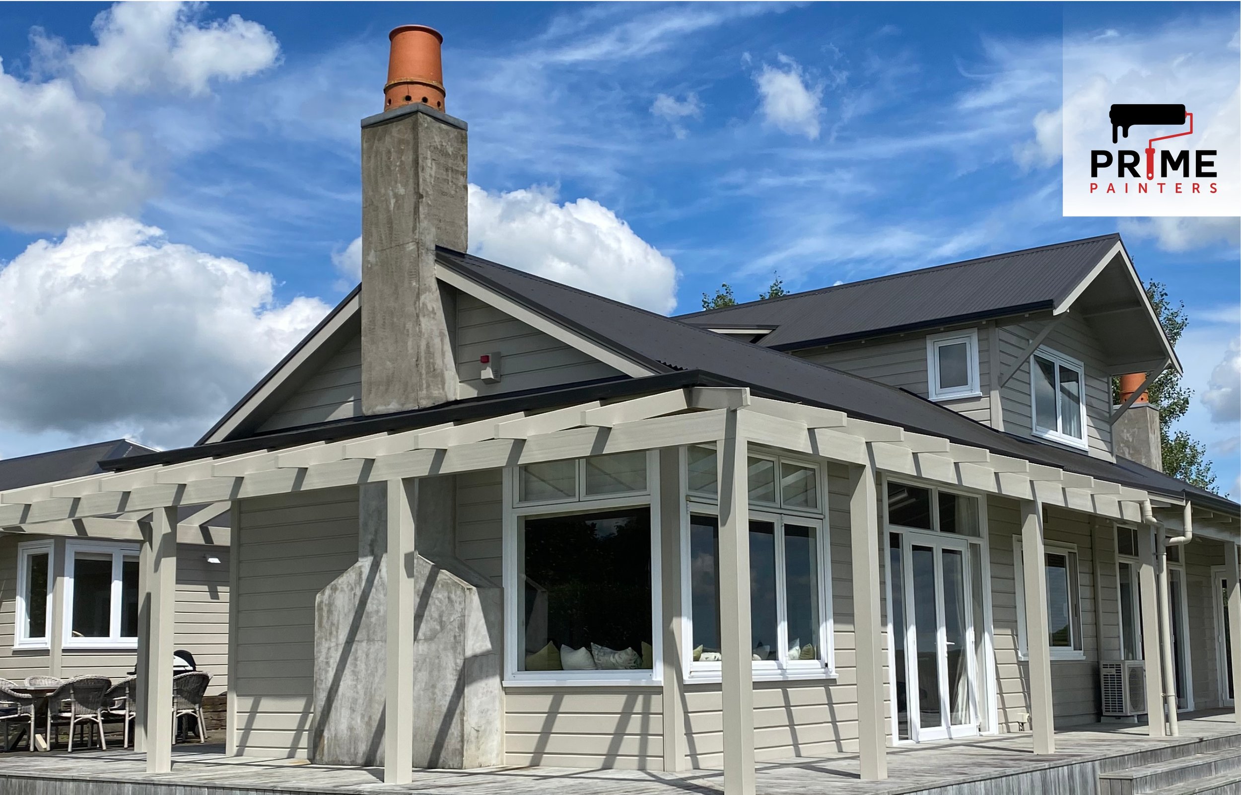 Exterior of a modern house with a deck, large windows, and a metal roof under a cloudy blue sky. The house has beige siding, and a prominent chimney with a concrete base and metal top. The Prime Painters logo is in the top right corner.