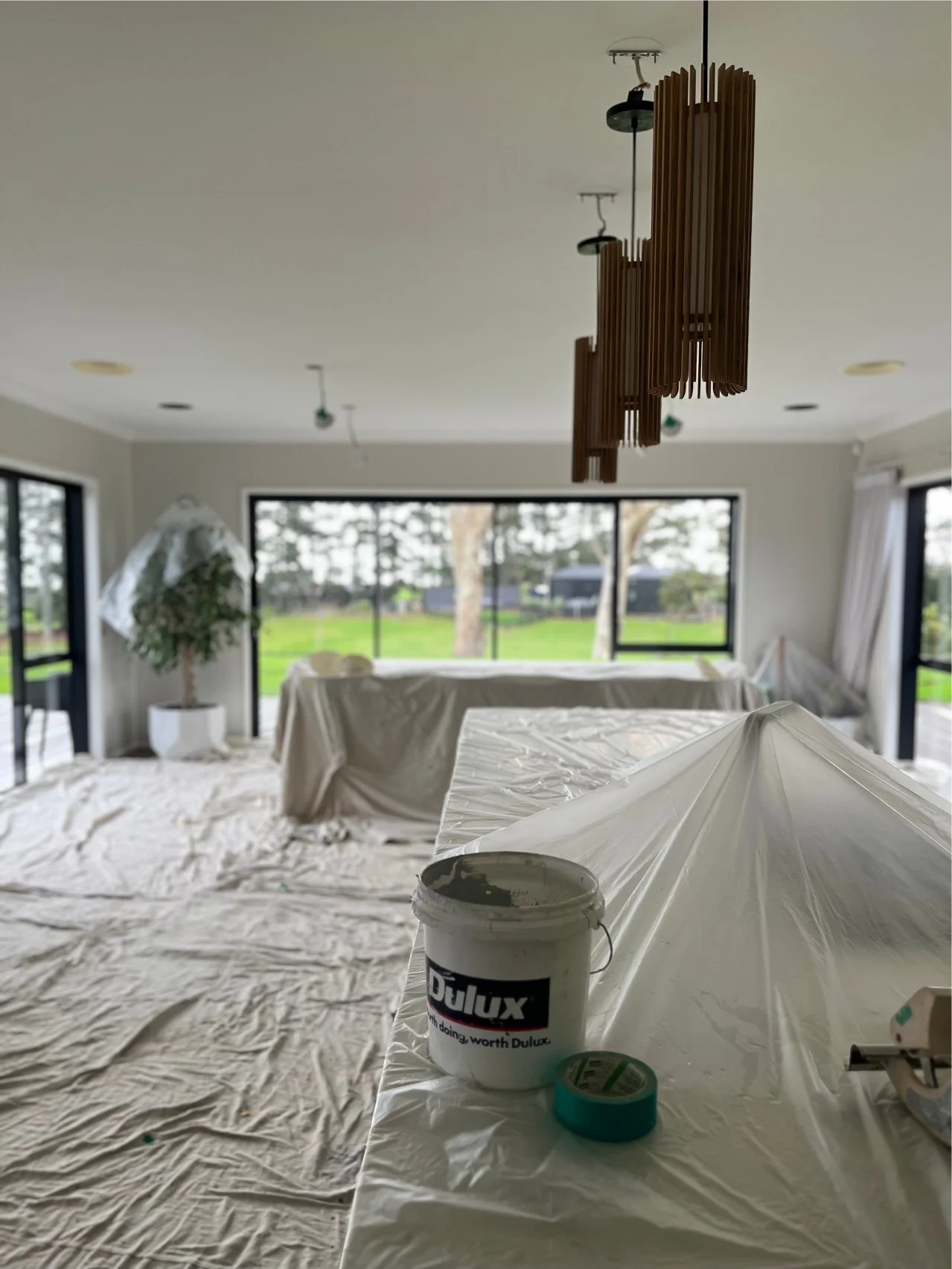 Living room under renovation with drop cloths on furniture and floor, a bucket of white paint, green painter's tape, and a ceiling light fixture hanging from the ceiling, large windows in the background revealing a green outdoor landscape.