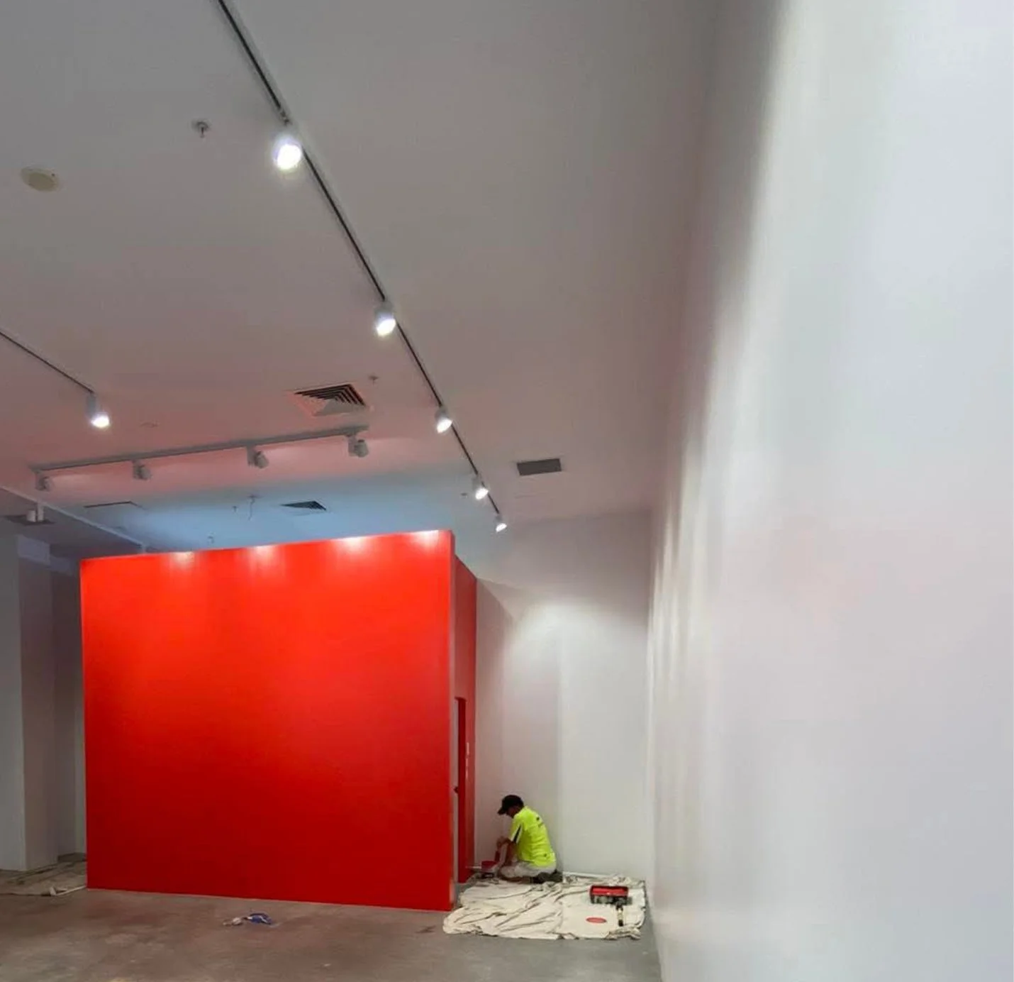 A worker in a neon yellow shirt and black cap assembling or painting the corner of a large bright red wall in an indoor space with white walls and ceiling. Ceiling lights and ventilation are visible.
