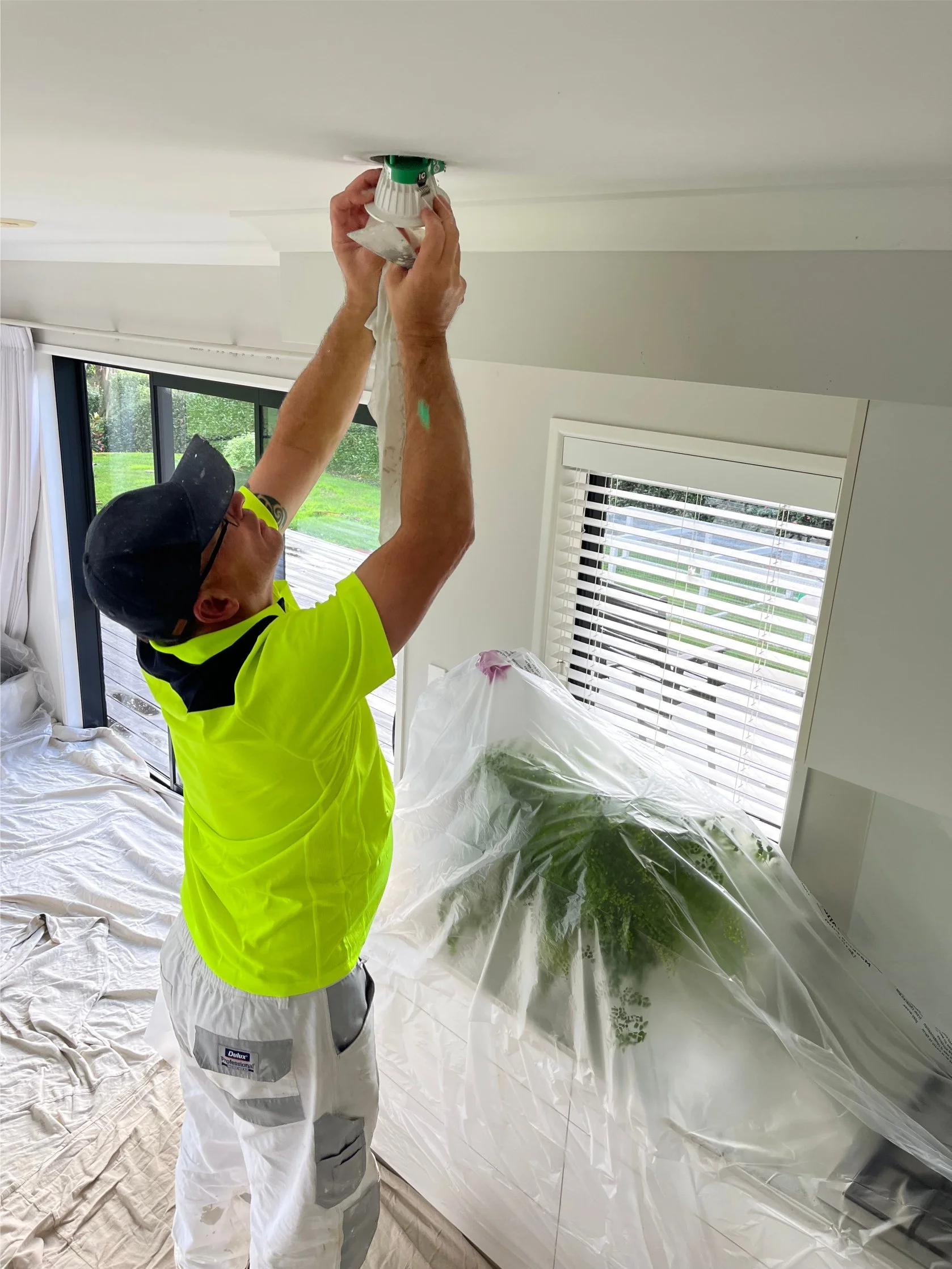 A man in a neon yellow shirt and white workpants removes light fitting on a ceiling in a bright room with large windows and a sliding glass door.