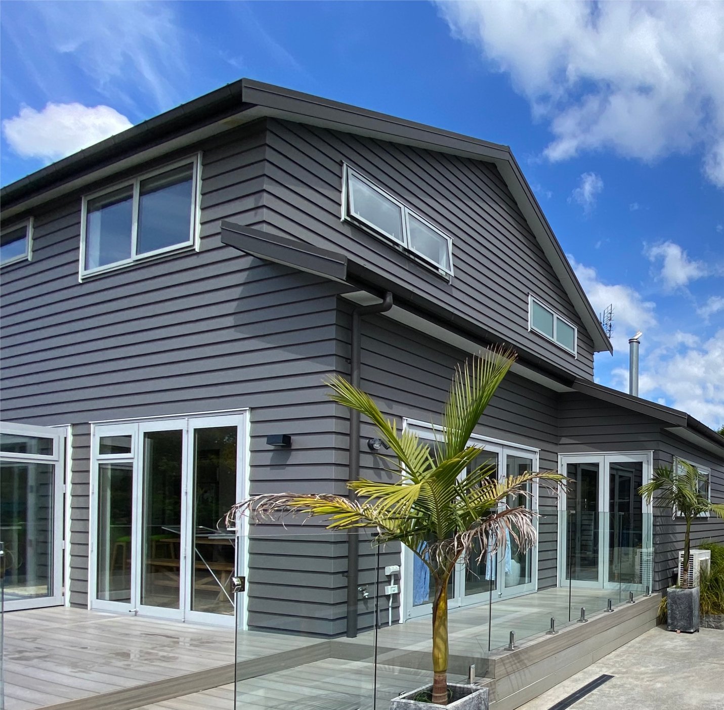 Modern gray house with large glass sliding doors and windows, surrounded by a glass railing, with small palm trees and a blue sky with white clouds.