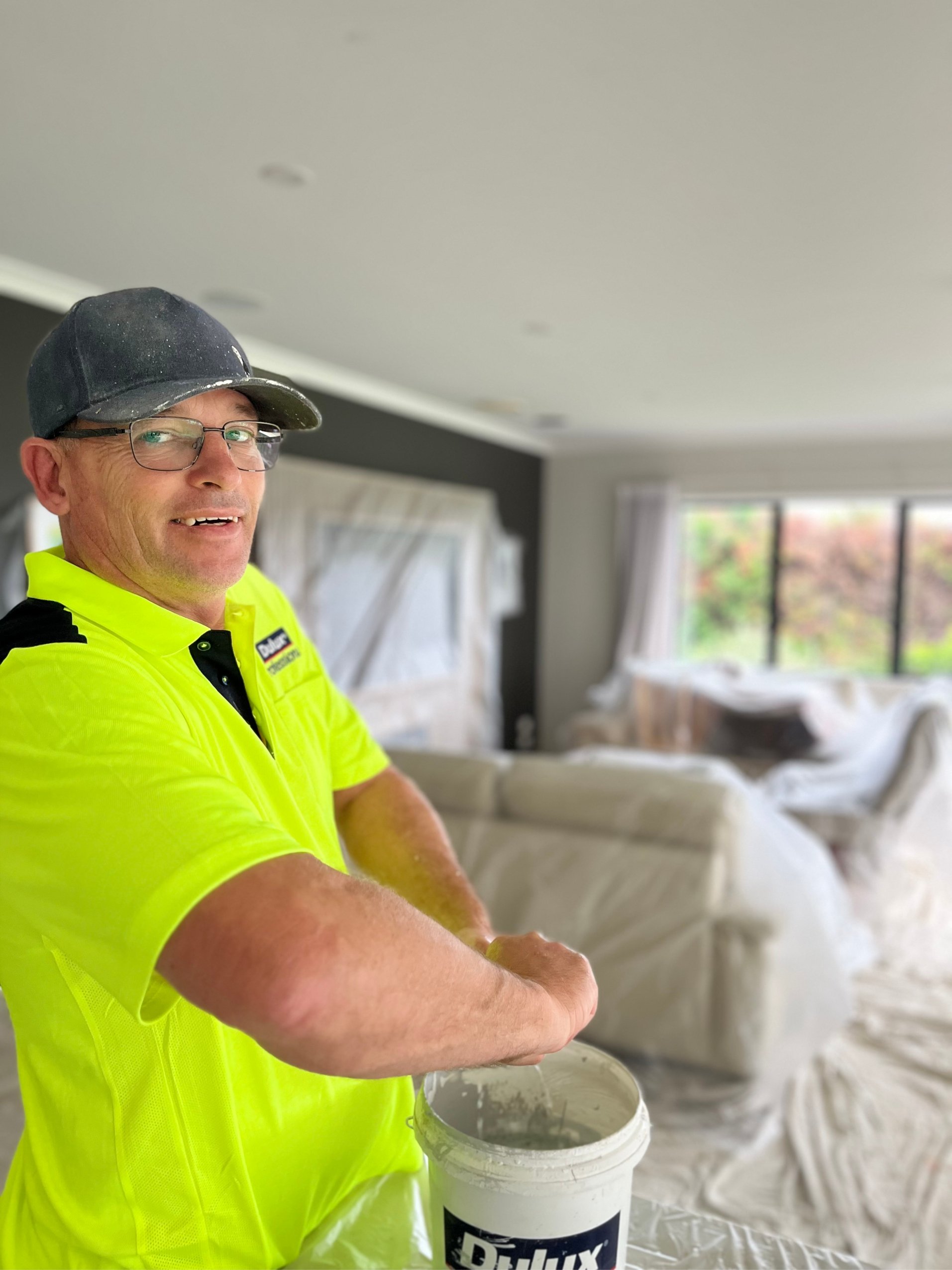 A man wearing a black cap, glasses, and a bright yellow safety shirt is working on a home renovation project inside a room. He is handling a bucket of white paint or primer, and the room appears to be covered in protective plastic, indicating ongoing painting or renovation work.