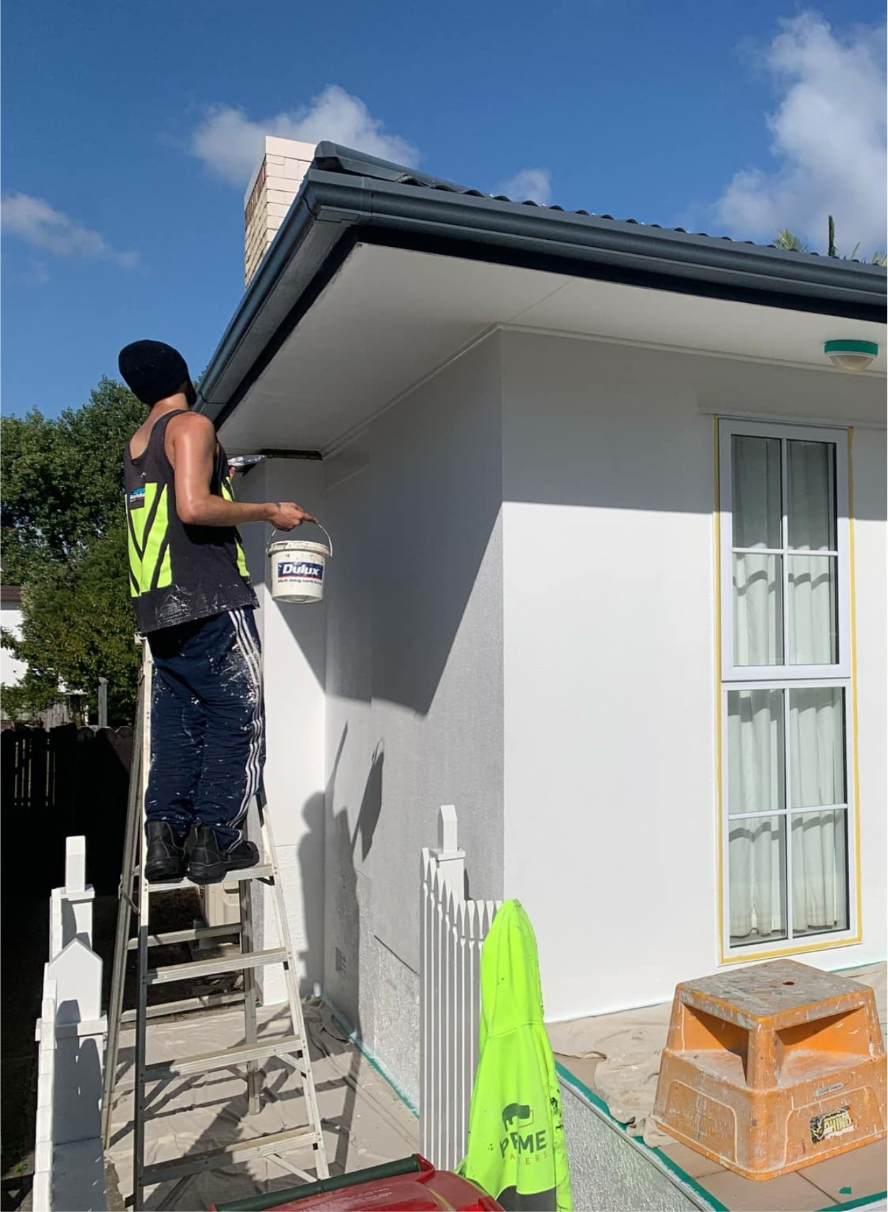 A person standing on a ladder painting the exterior wall of a house with white paint. The person is wearing a black beanie, a black sleeveless shirt with yellow reflective stripes, and blue pants with white stripes. They are holding a paintbrush in one hand and a bucket of paint in the other. The house has a window with white curtains and yellow trim, a small fence, and construction materials on the ground.