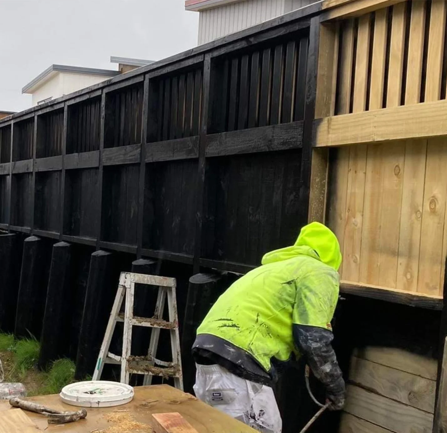 A worker in a yellow safety jacket and white pants is working on a wooden fence installation, using a spray gun. Fence has black stained wood on one side and natural light-colored wood on the other. A ladder and a work table with tools are nearby.