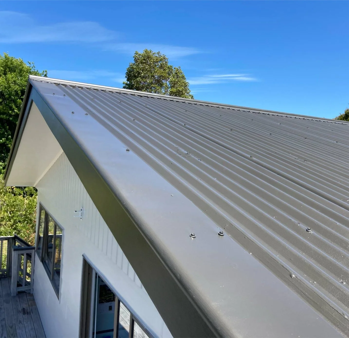 Close-up of a metal roof with screws on a white house, under a blue sky with trees in the background.