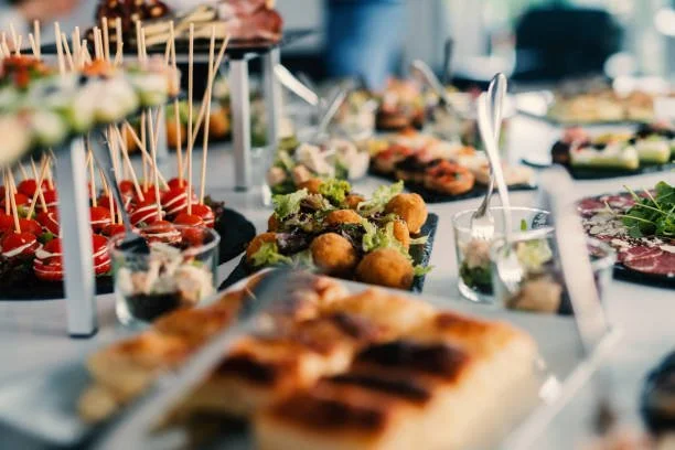 Assorted appetizers and finger foods on a buffet table, including skewers with cherry tomatoes and cheese, mini sandwiches, croquettes, and sliced cured meats.