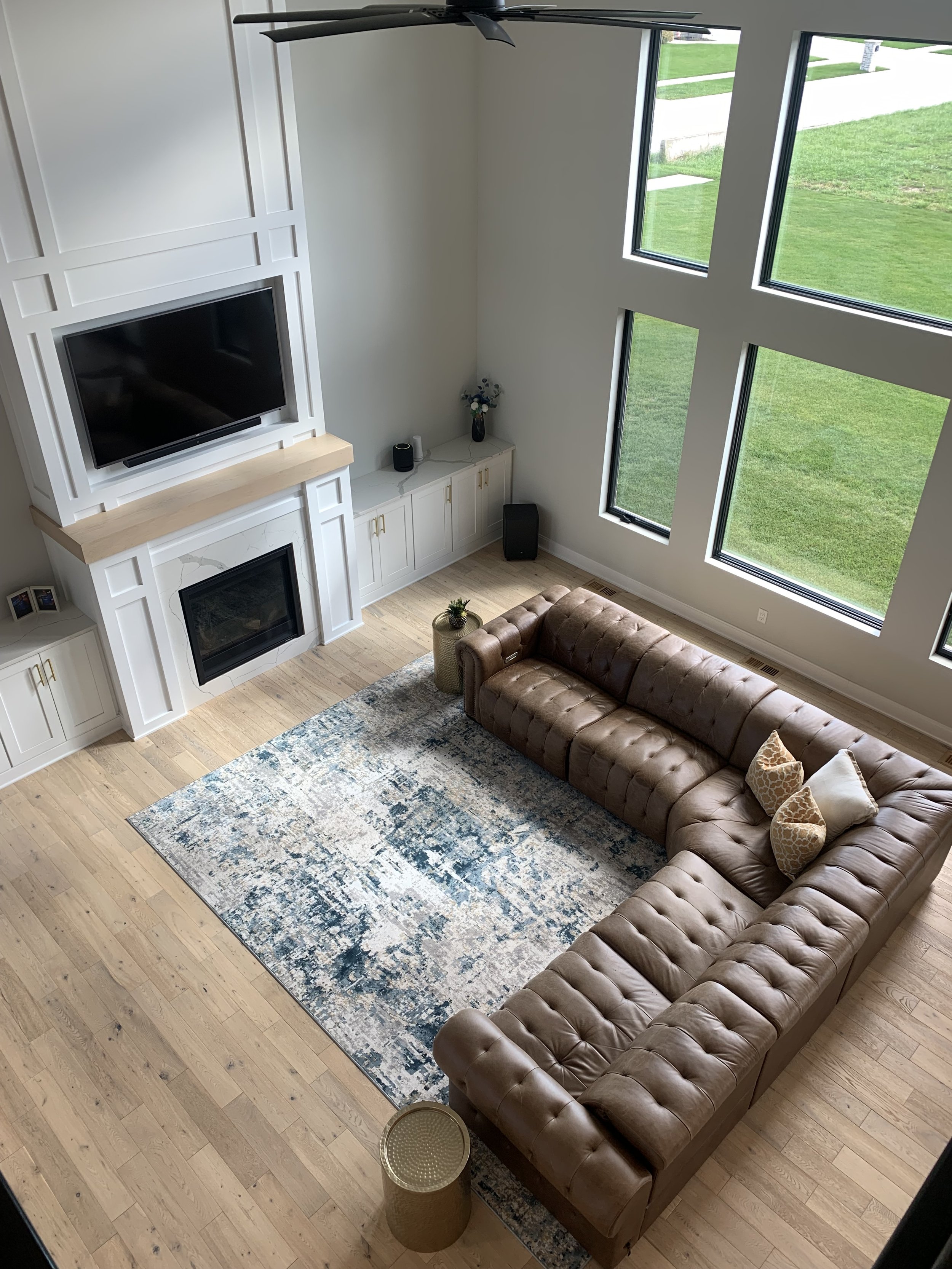 Living room with large leather sectional, area rug, fireplace, mounted TV, large windows, and light wood flooring.