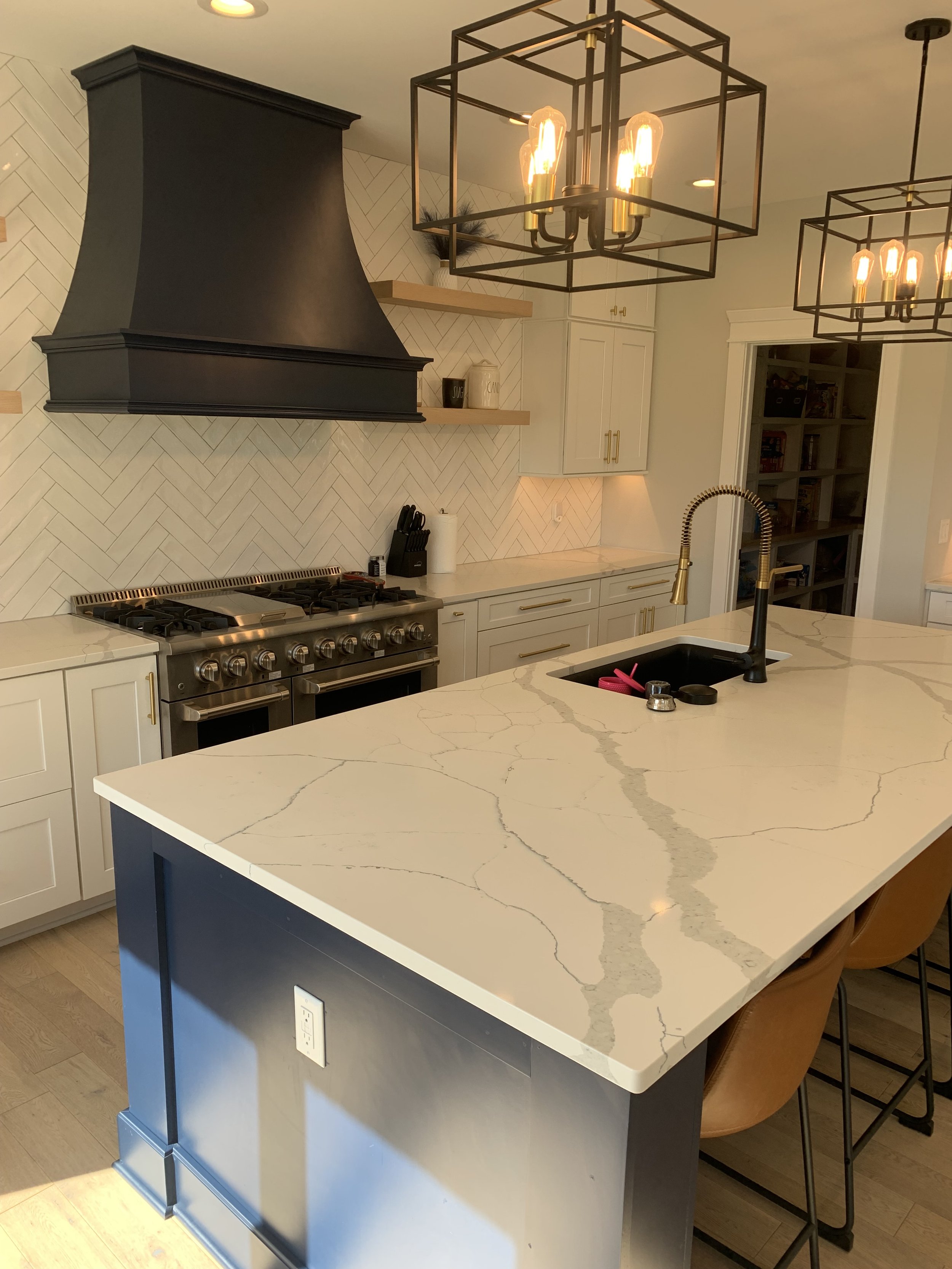 Modern kitchen with a large white marble island, black sink, gold faucet, black stove, black range hood, open shelving, white cabinetry, and black geometric pendant lights.