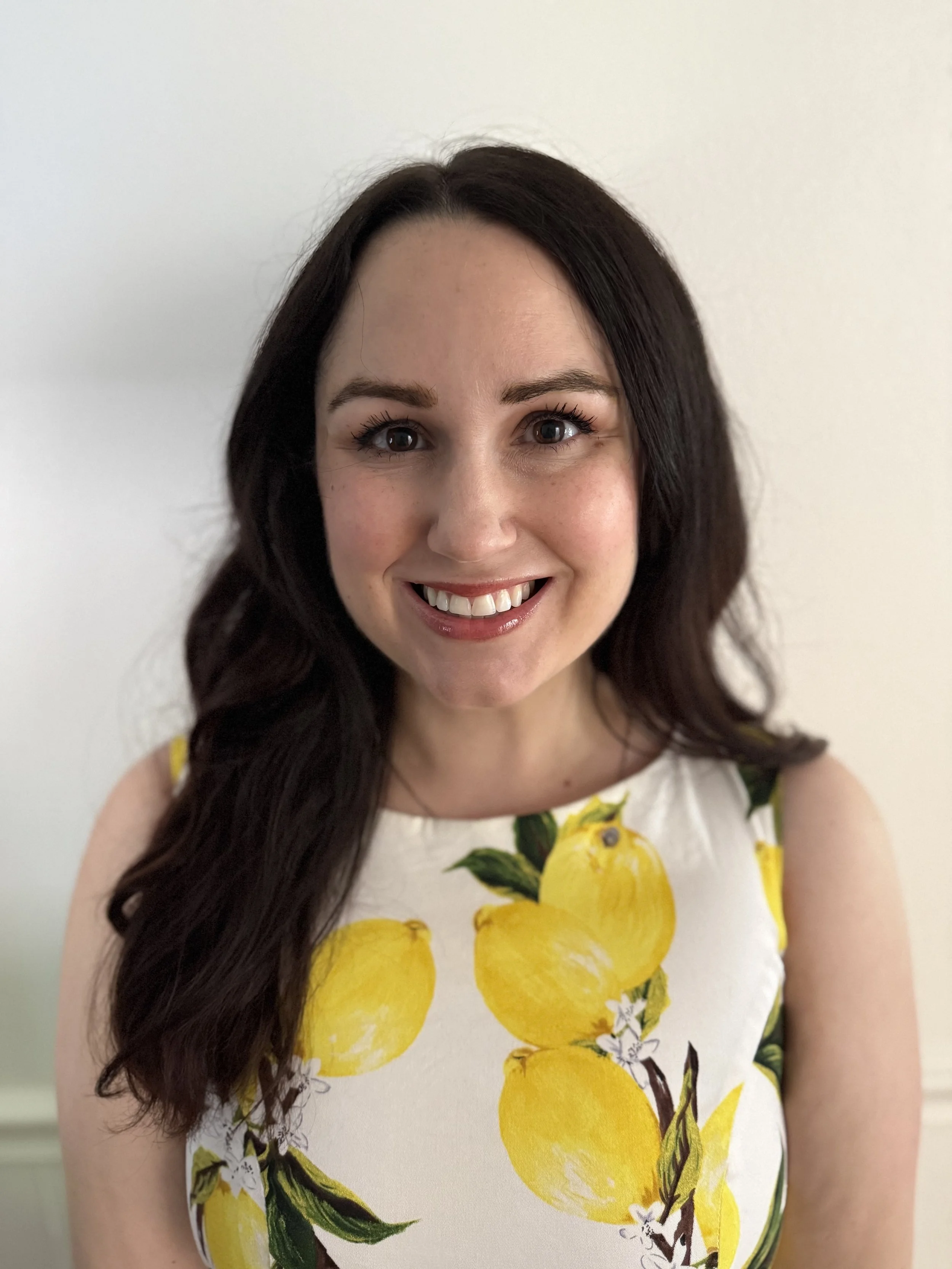 A woman with long dark hair, smiling, wearing a sleeveless dress with a lemon print, standing against a plain white wall.