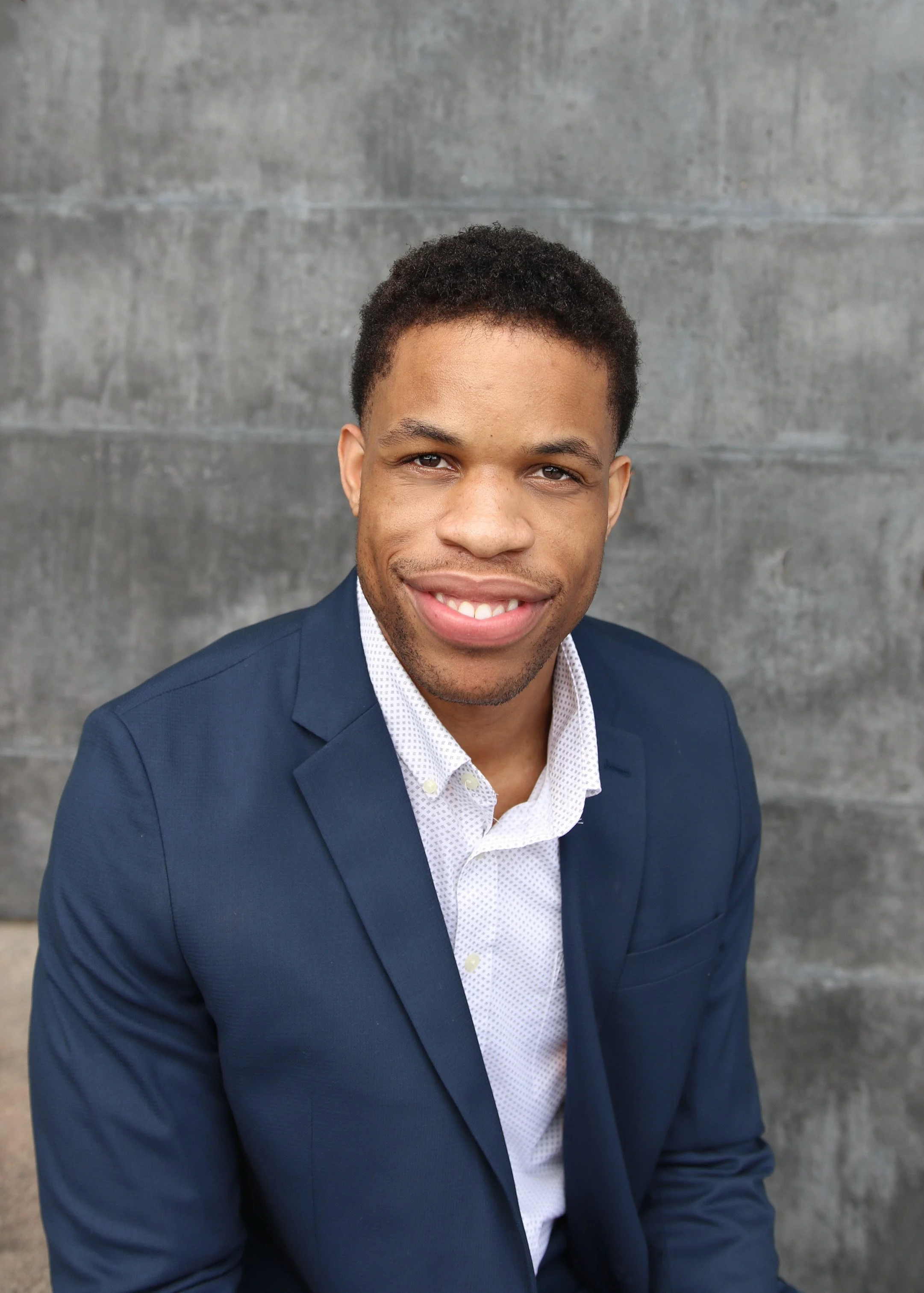 A smiling man in a navy blue suit and white patterned shirt posing outdoors against a concrete wall.