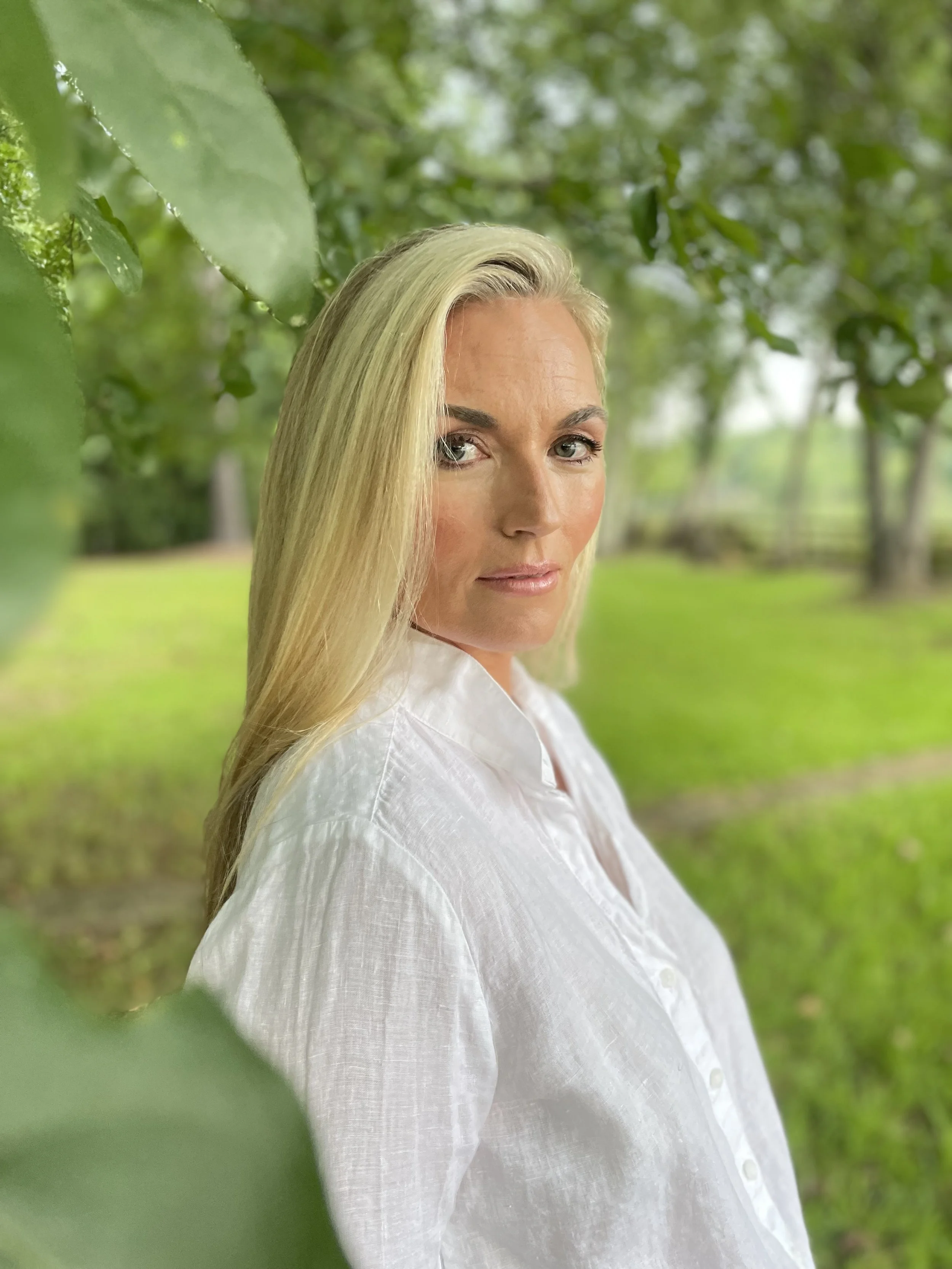 A woman with long blonde hair and fair skin standing outdoors among green trees and foliage, wearing a white buttoned shirt.