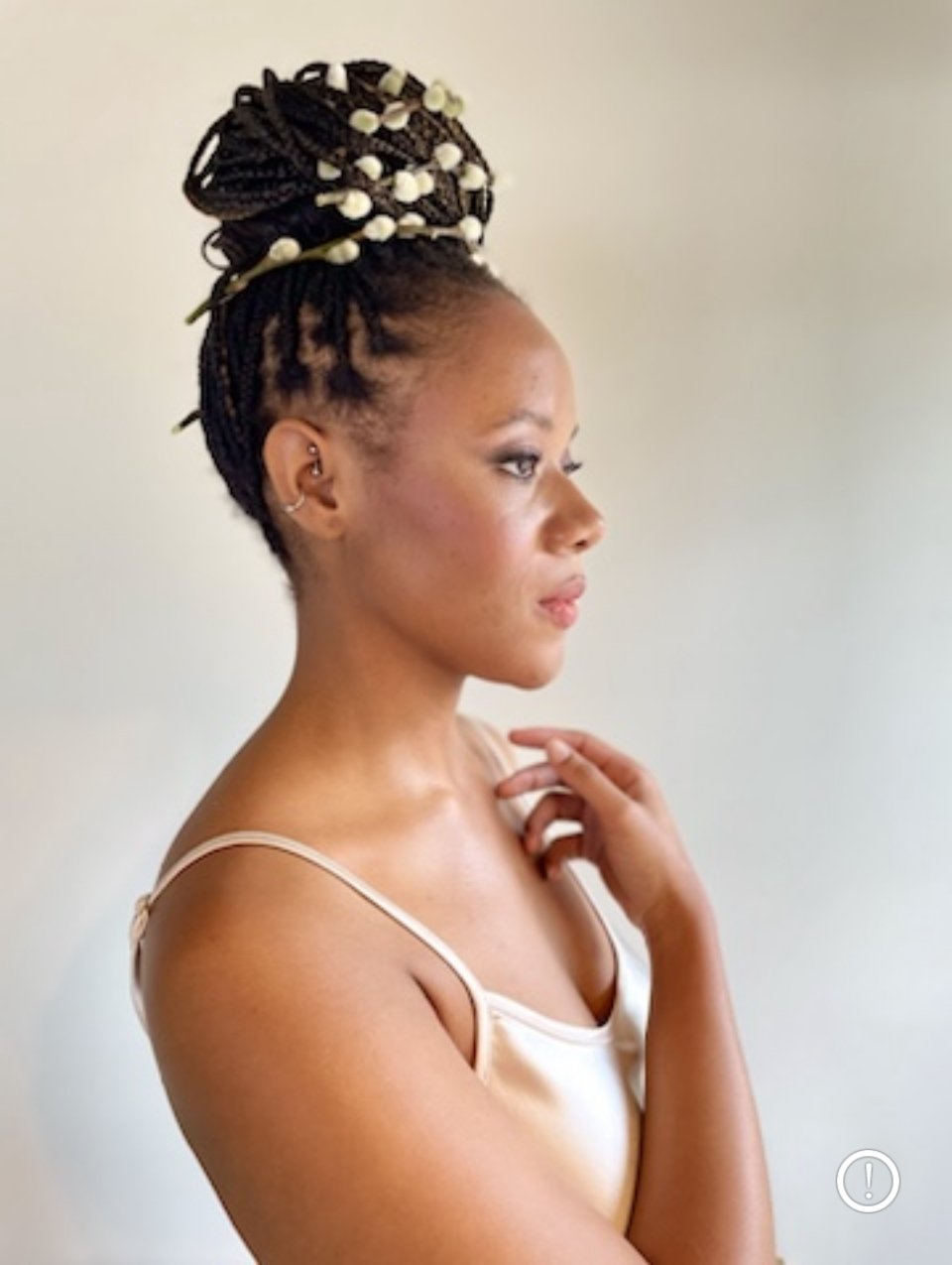Side profile of a woman with her hair styled in long black braids with white beads, wearing a cream-colored camisole top, against a plain neutral background.