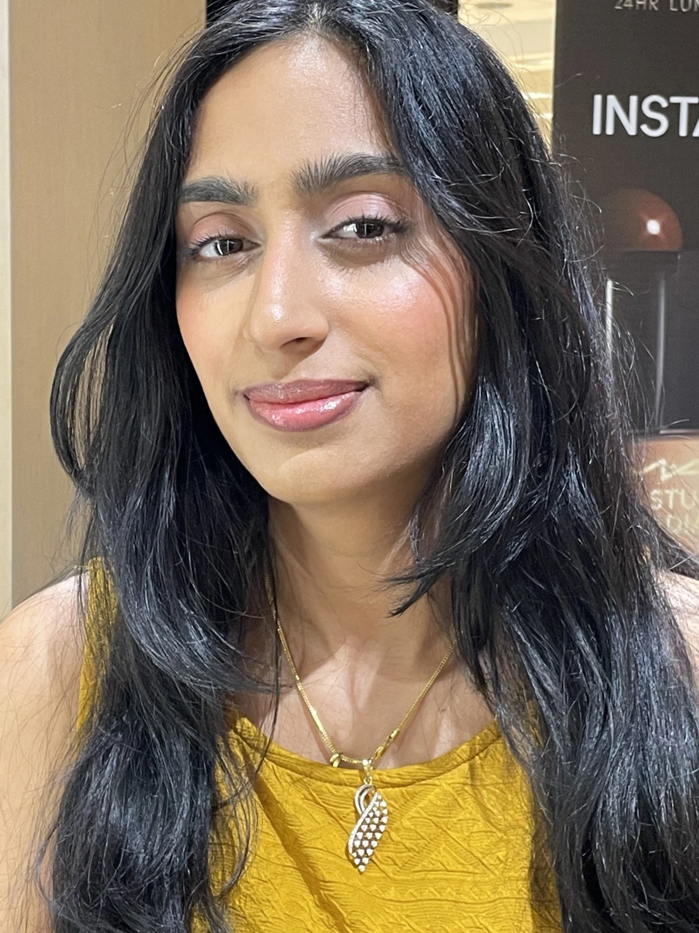 Close-up of a woman with long dark hair, wearing a yellow top and gold necklace, smiling slightly, in an indoor setting.