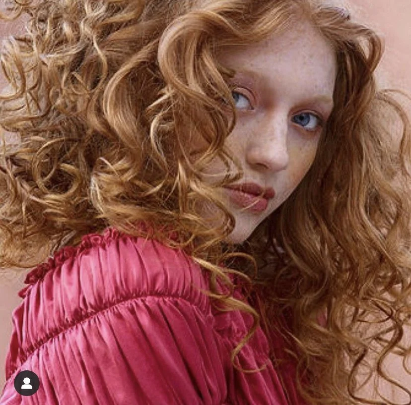 Close-up of a woman with curly red hair, blue eyes, and freckles, wearing a pink pleated top.