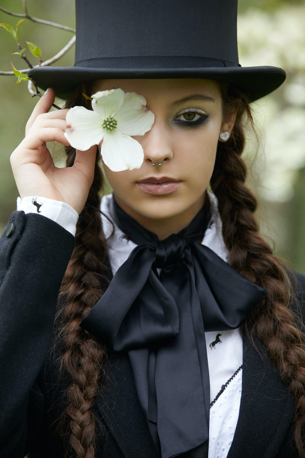 Young woman with braided hair, wearing a black top hat, black blazer, white shirt, and black bow tie, holding a white flower over her eye outdoors.