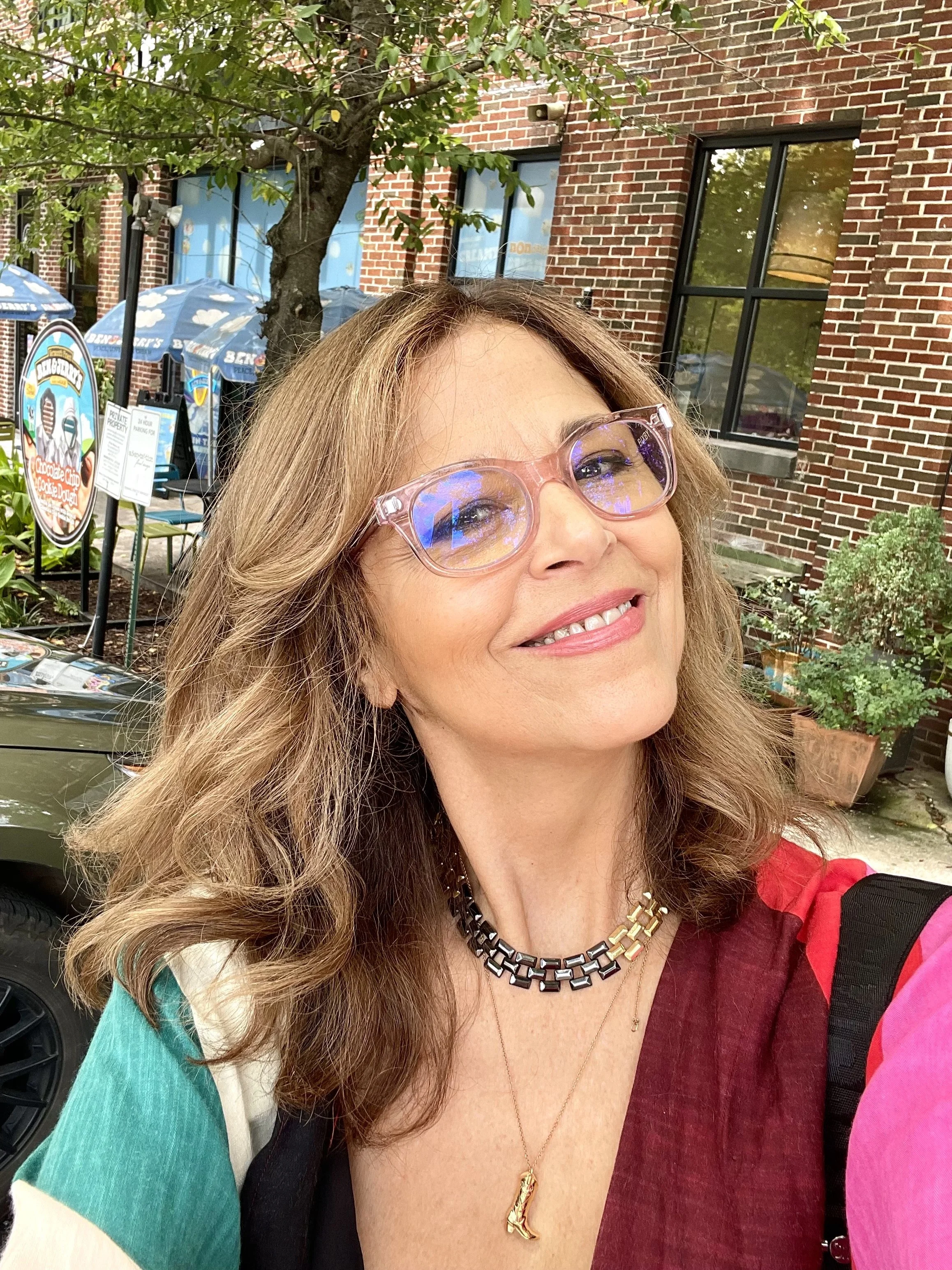 A woman with wavy, shoulder-length hair, wearing glasses with pink frames, smiling, and dressed in a multi-colored top with shades of red, green, cream, and pink. She has a black and gold necklace and a gold necklace with a letter 'L'. She is outdoors with a brick building, trees, and outdoor seating with umbrellas in the background.