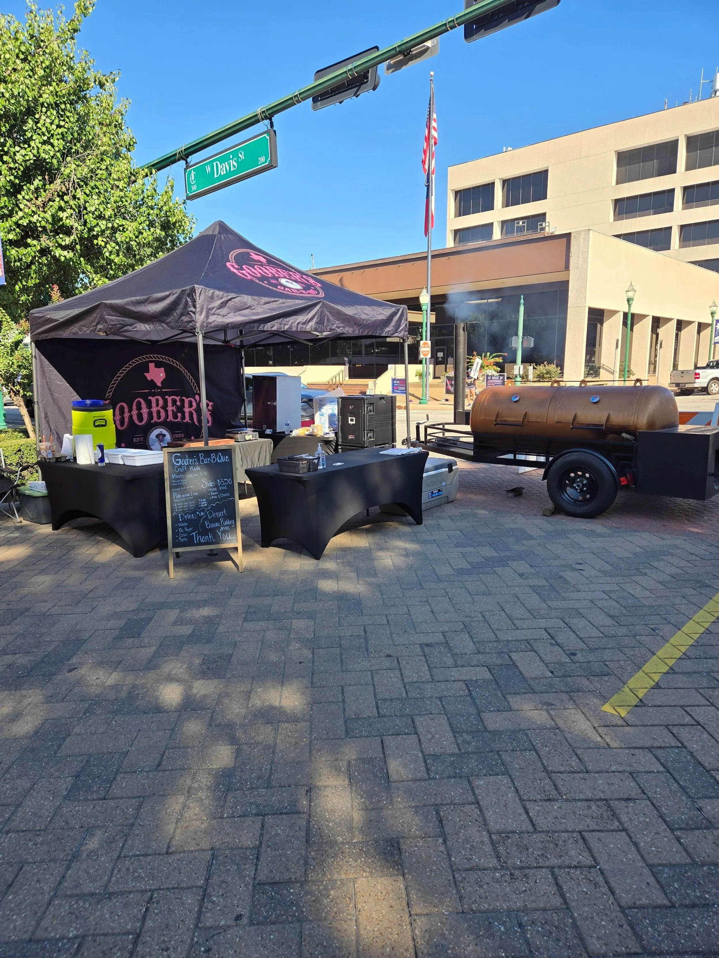 A barbecue food stall with a black canopy and a menu board, set up on a city sidewalk with a large barbecue smoker on a trailer nearby. The background shows a building, street signs, trees, and the American flag.