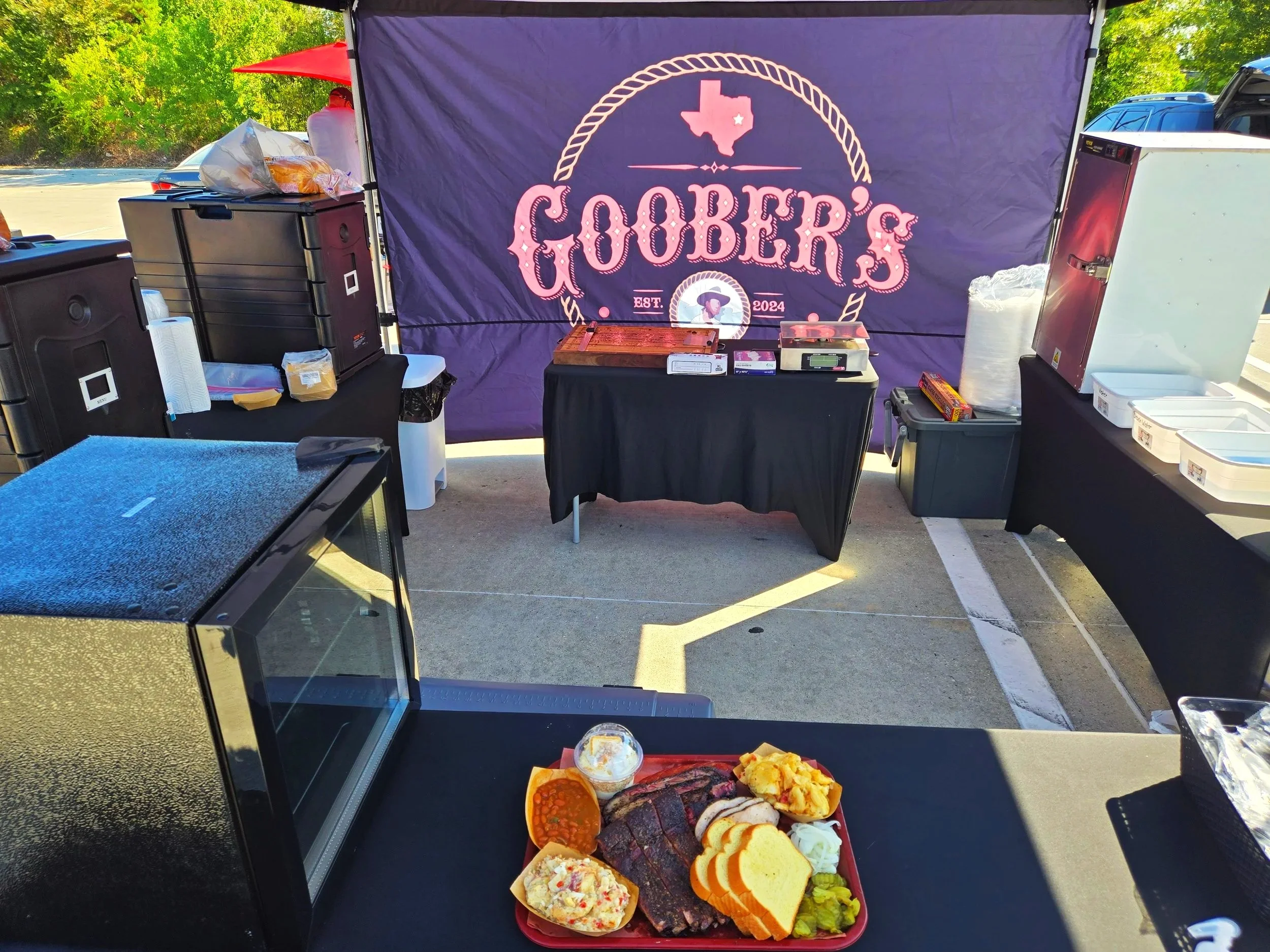 A table with sliced bread, fried foods, and a cup of dip or sauce in the foreground, with a food stall in the background featuring a purple banner with the text "Goober's" and an illustration of Texas.