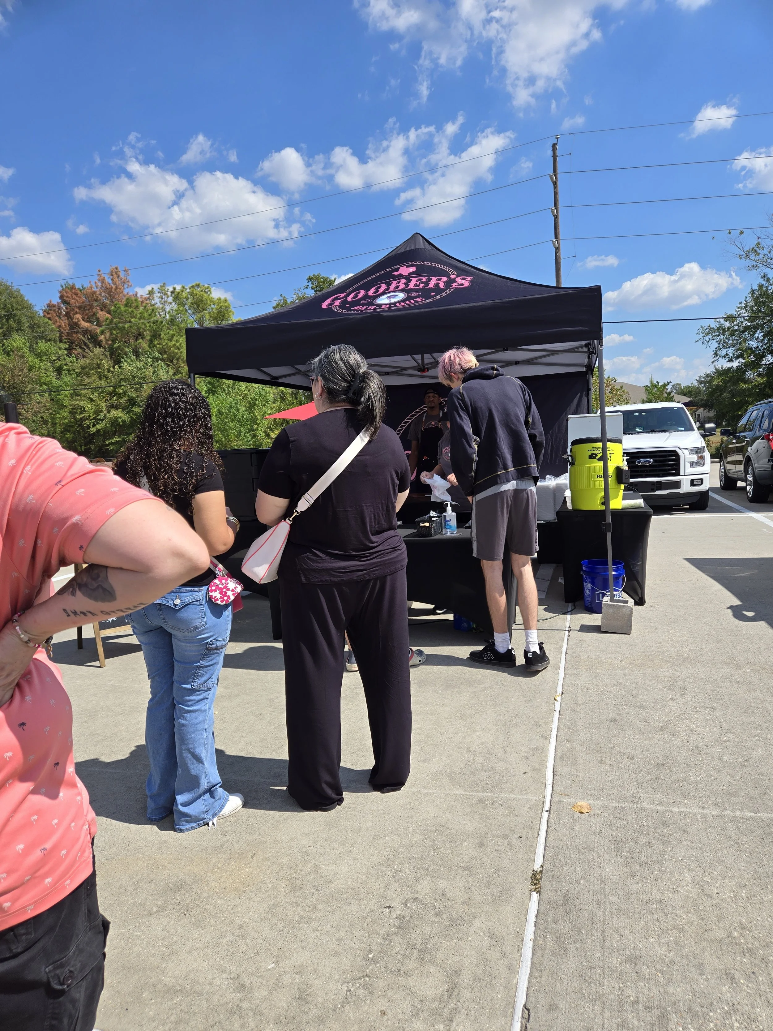 People waiting in line at a food truck with a black tent that has pink lettering, in a parking lot on a sunny day with blue sky and scattered clouds.