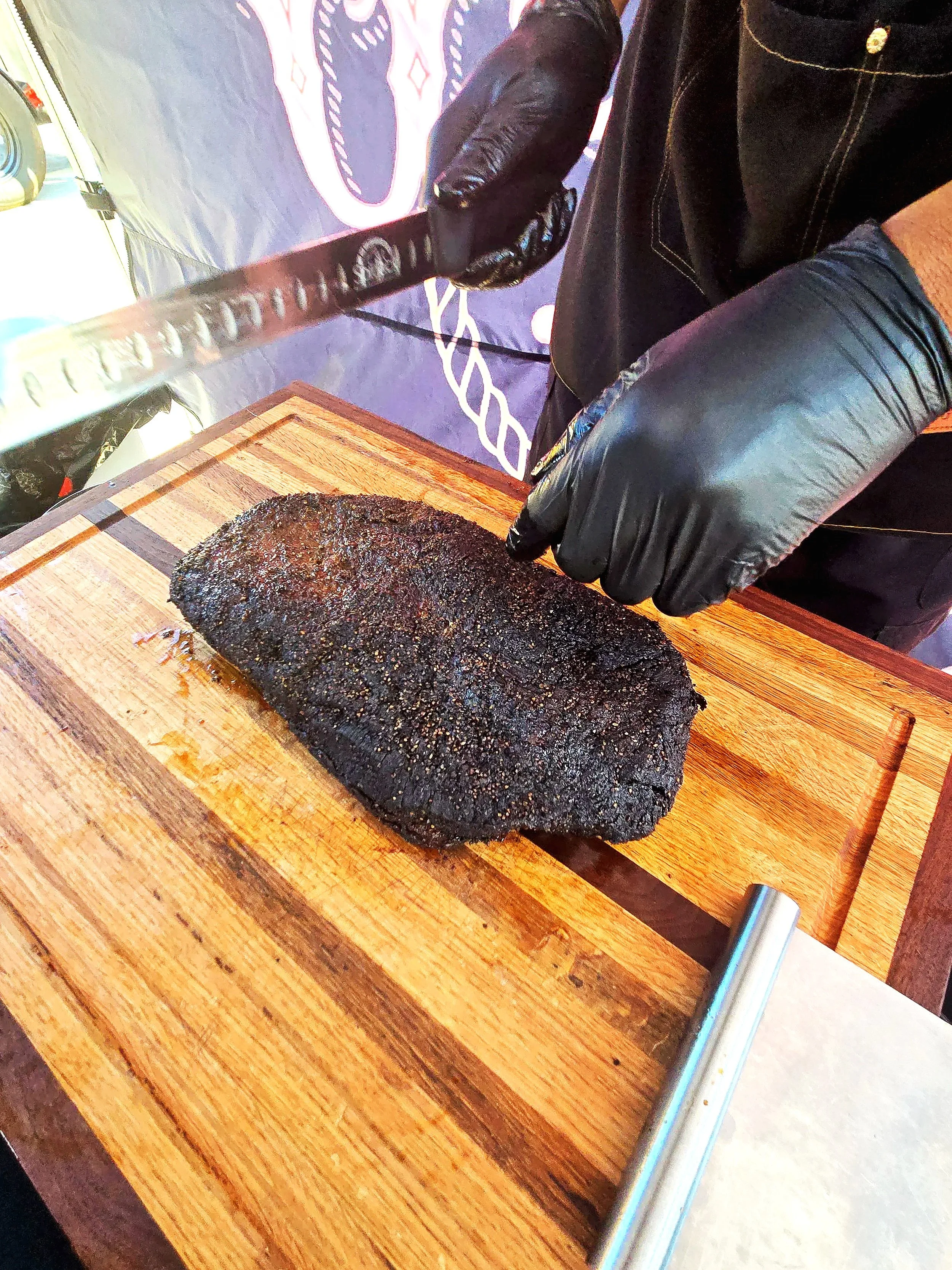 Person in black gloves slicing a smoked brisket on a wooden cutting board with a large knife.