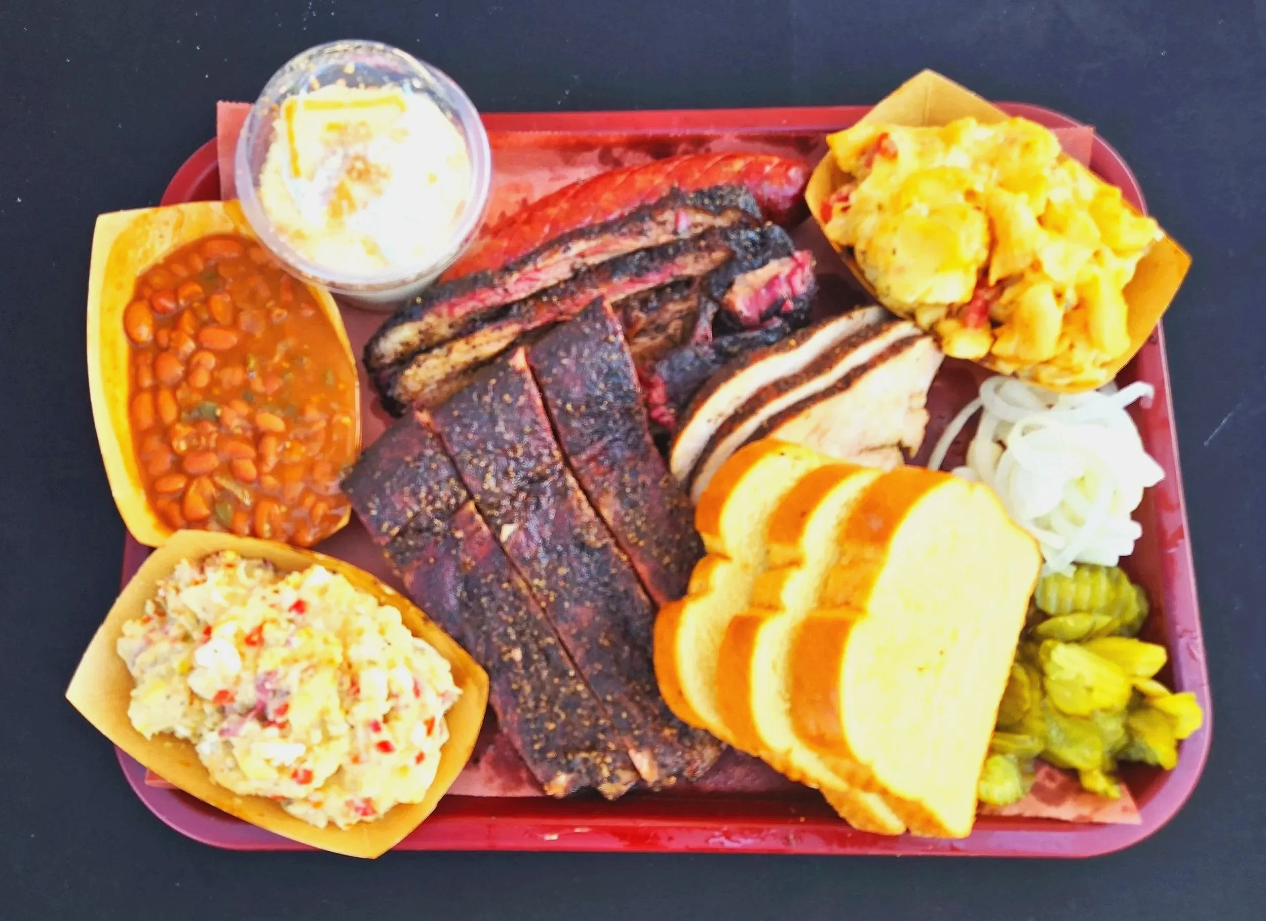 Barbecue platter with sliced brisket, sausage, baked beans, potato salad, scrambled eggs, sliced bread, pickles, and coleslaw.