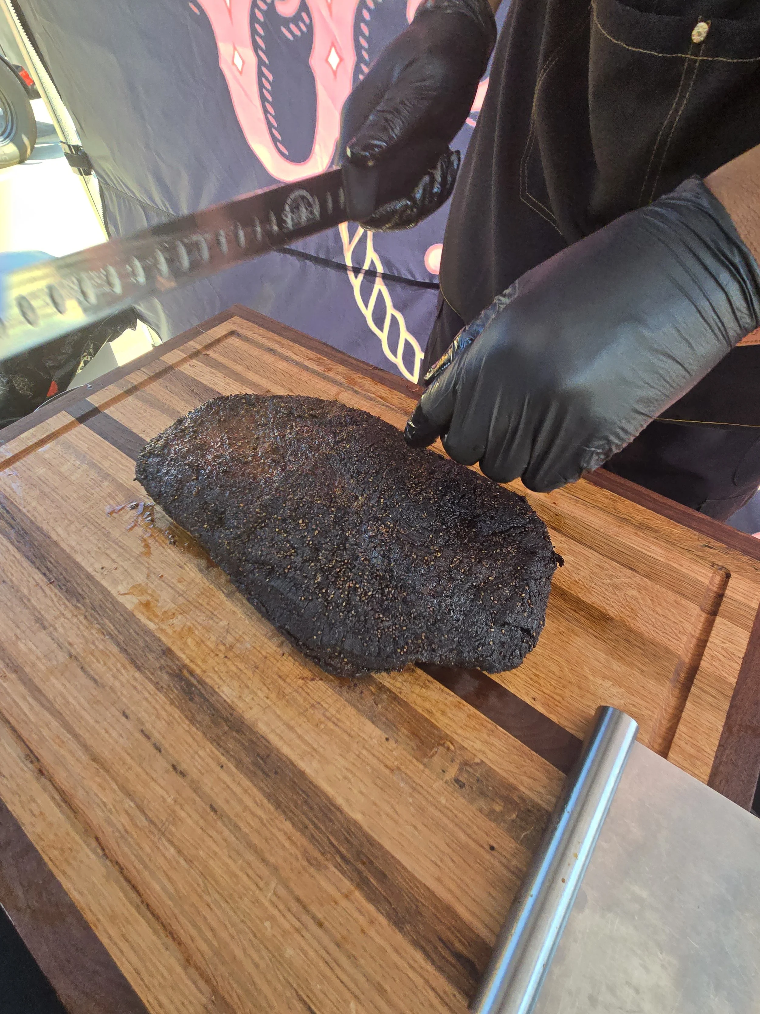 A person wearing black gloves slicing a large, cooked piece of meat on a wooden cutting board.