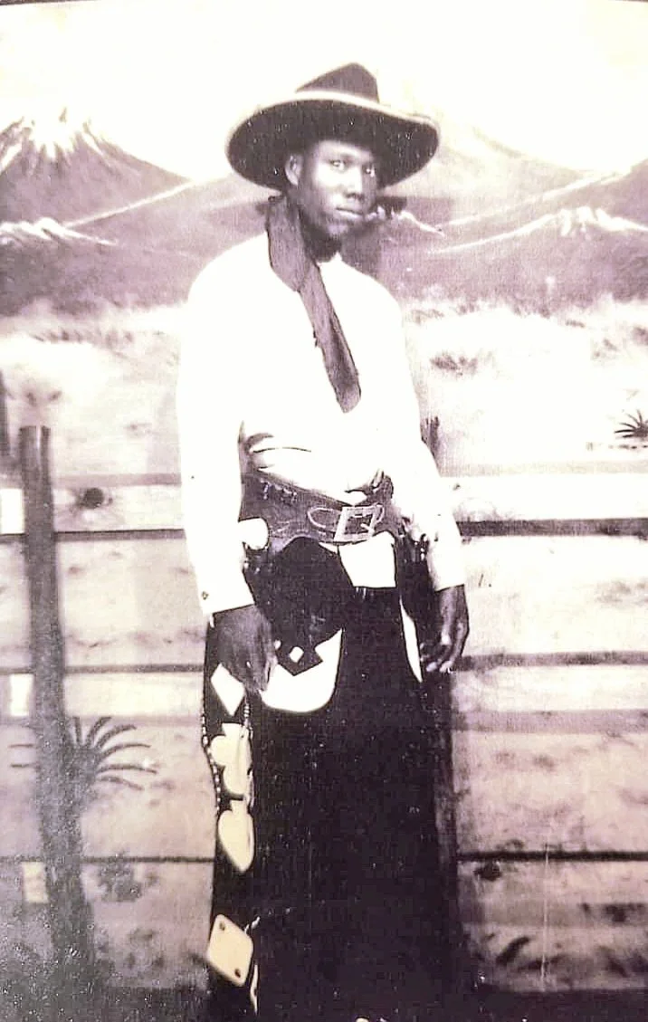 A Black man dressed as a cowboy, standing with arms crossed, wearing a wide-brimmed hat, in front of a painted mountain and mountain range background.