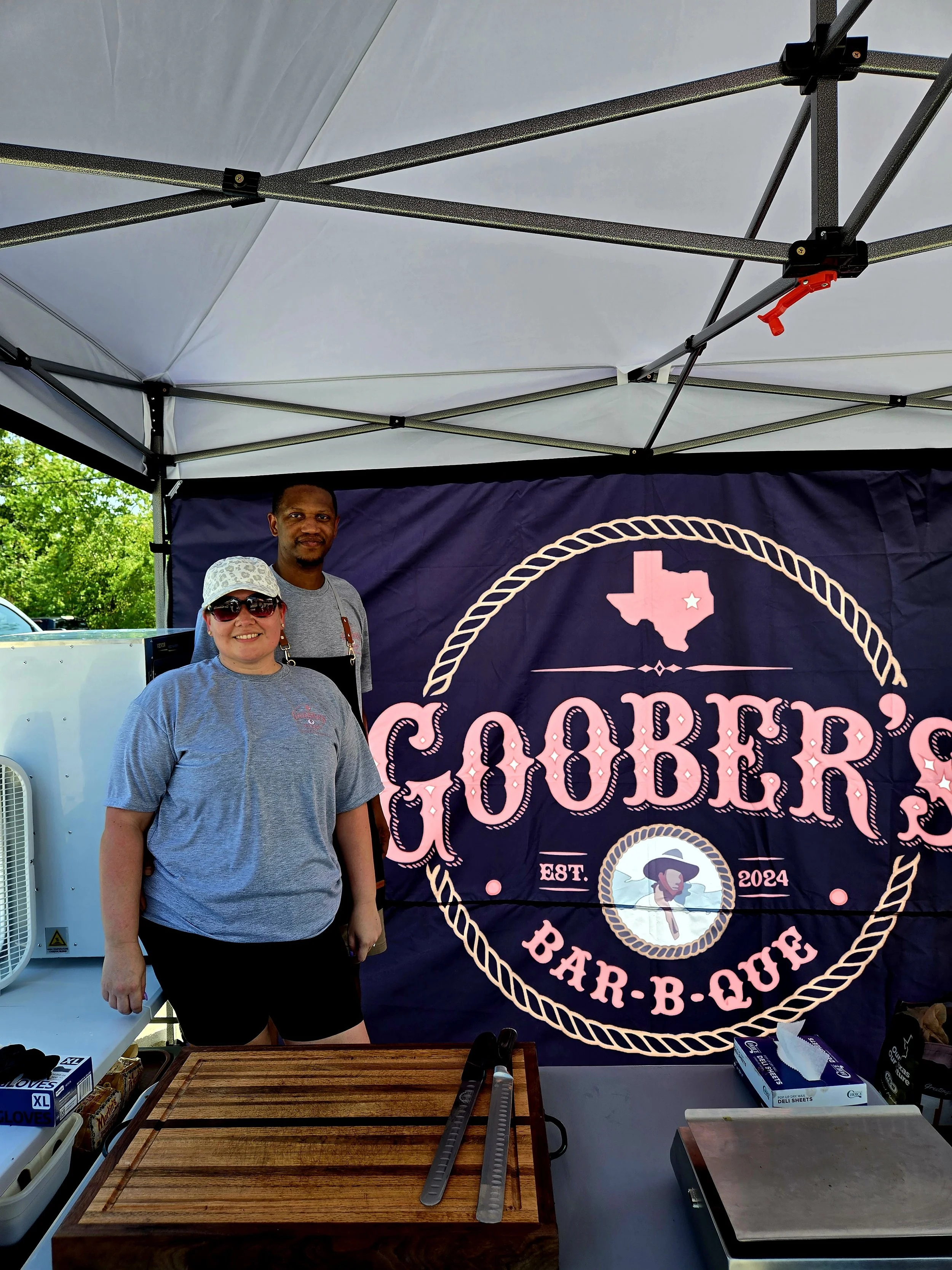 Two women standing in front of a Goober's Bar-B-Que banner inside a tent, with a wooden cutting board and grilling tongs on a table in front.