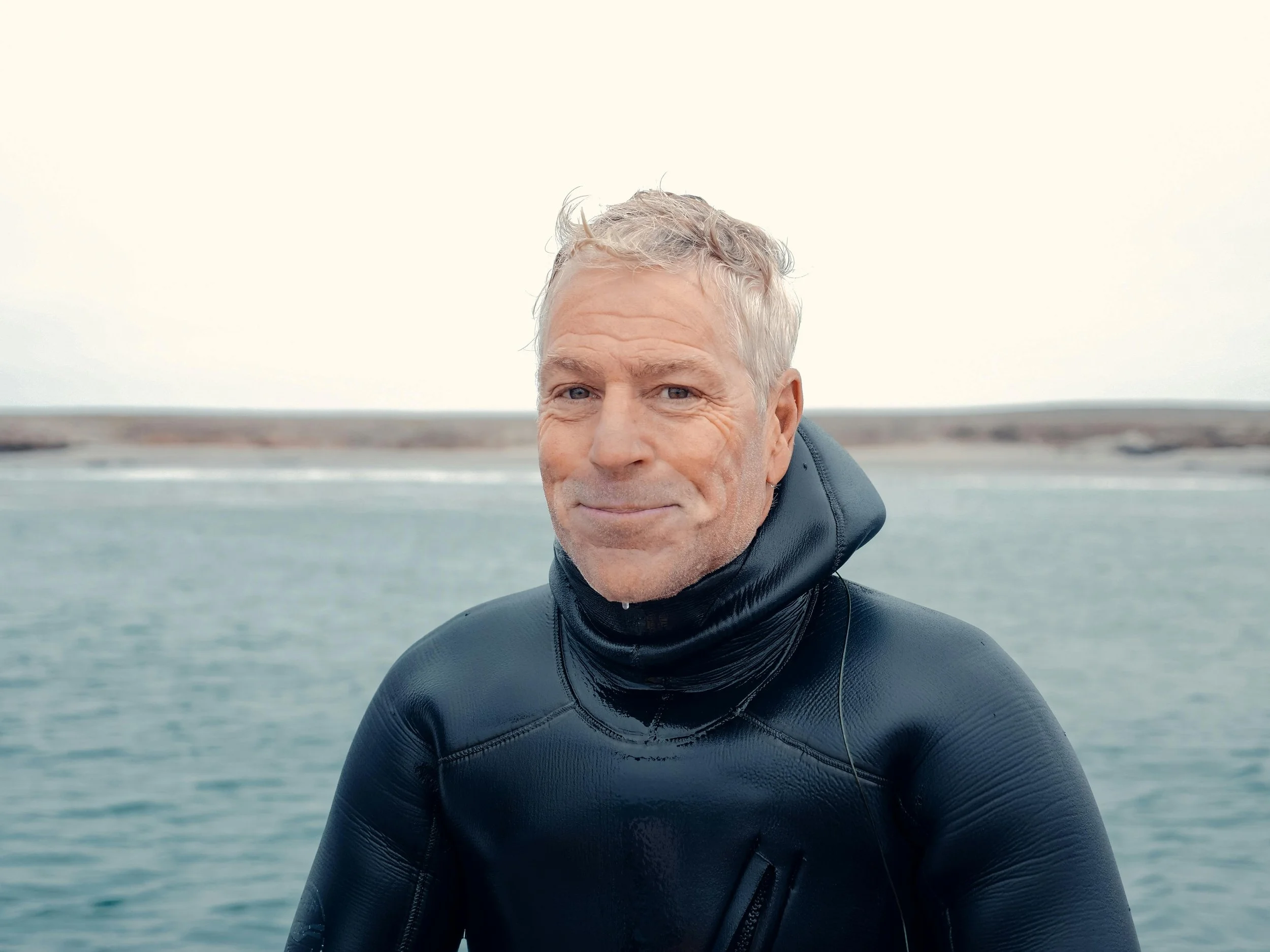 A senior man in a black wetsuit standing near a body of water, with a distant shoreline, under a cloudy sky.