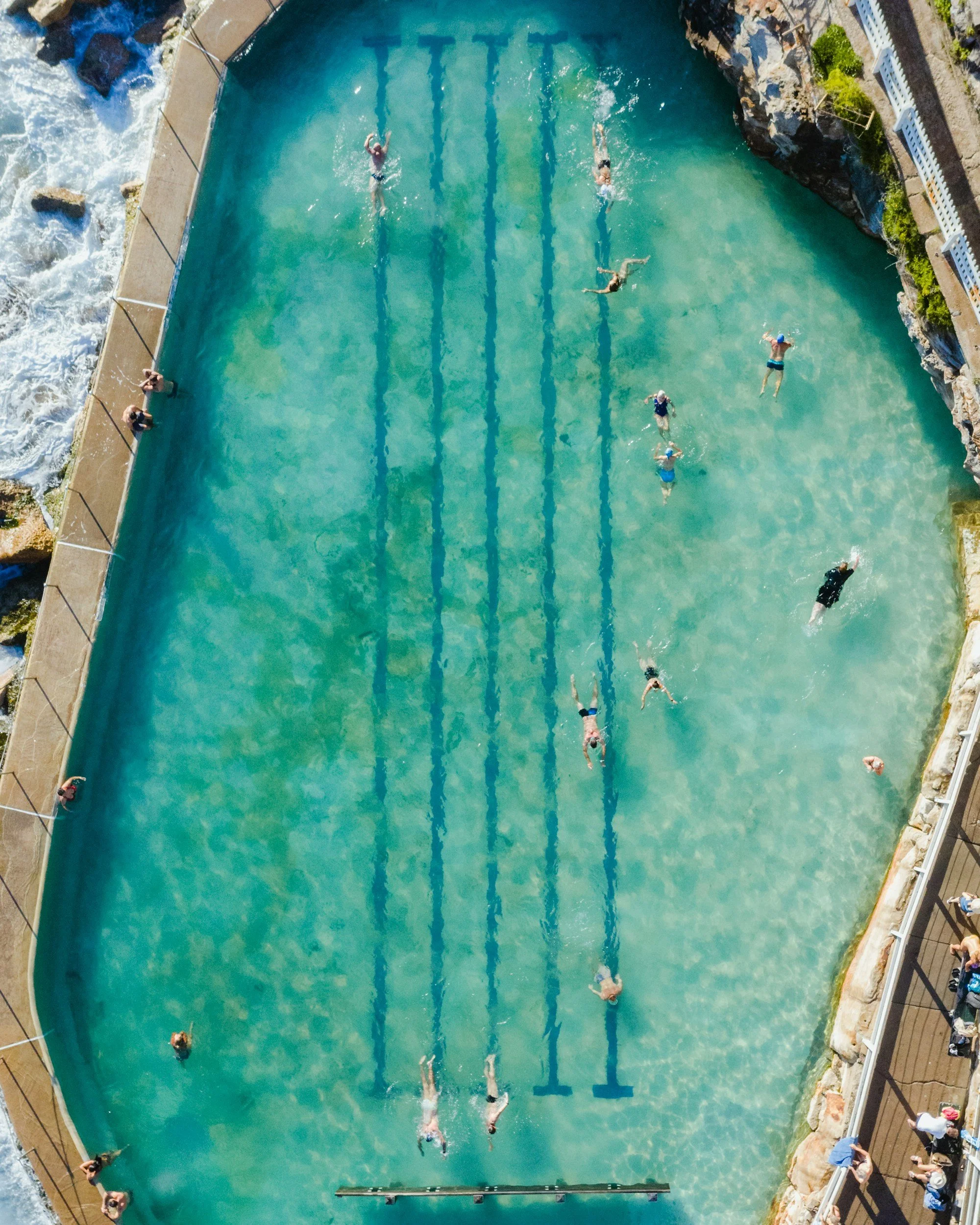 People swimming in an outdoor pool with lanes, surrounded by a railing and rocky landscape.