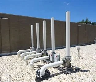 Series of white pipes and valves on gravel ground against a brown wall and blue sky.