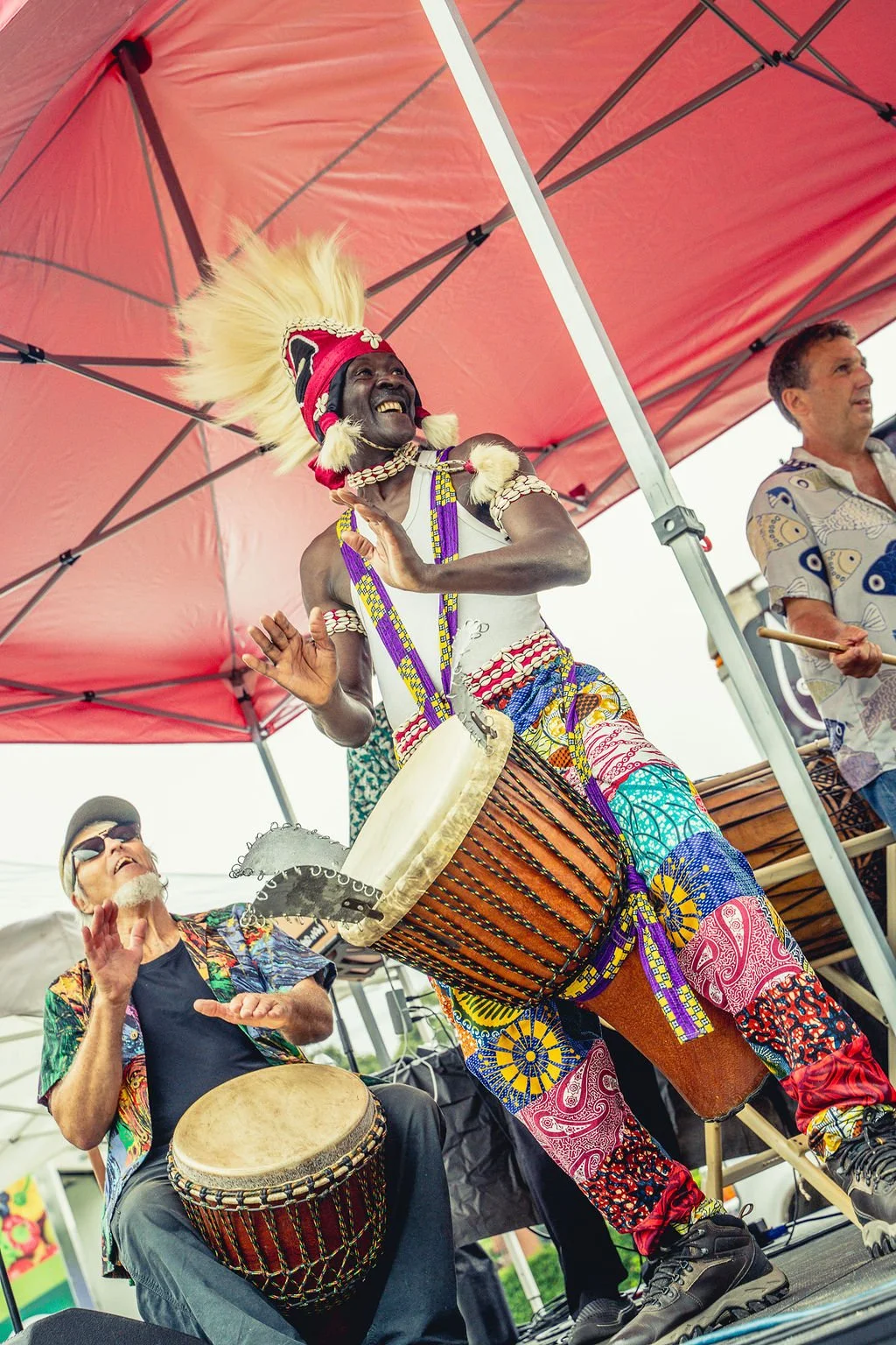 Musicians performing on stage at New England BIPOC Festival under a red canopy, including a smiling man with dark skin and a large feathered headdress playing a drum, and a man with light skin and sunglasses playing a drum.